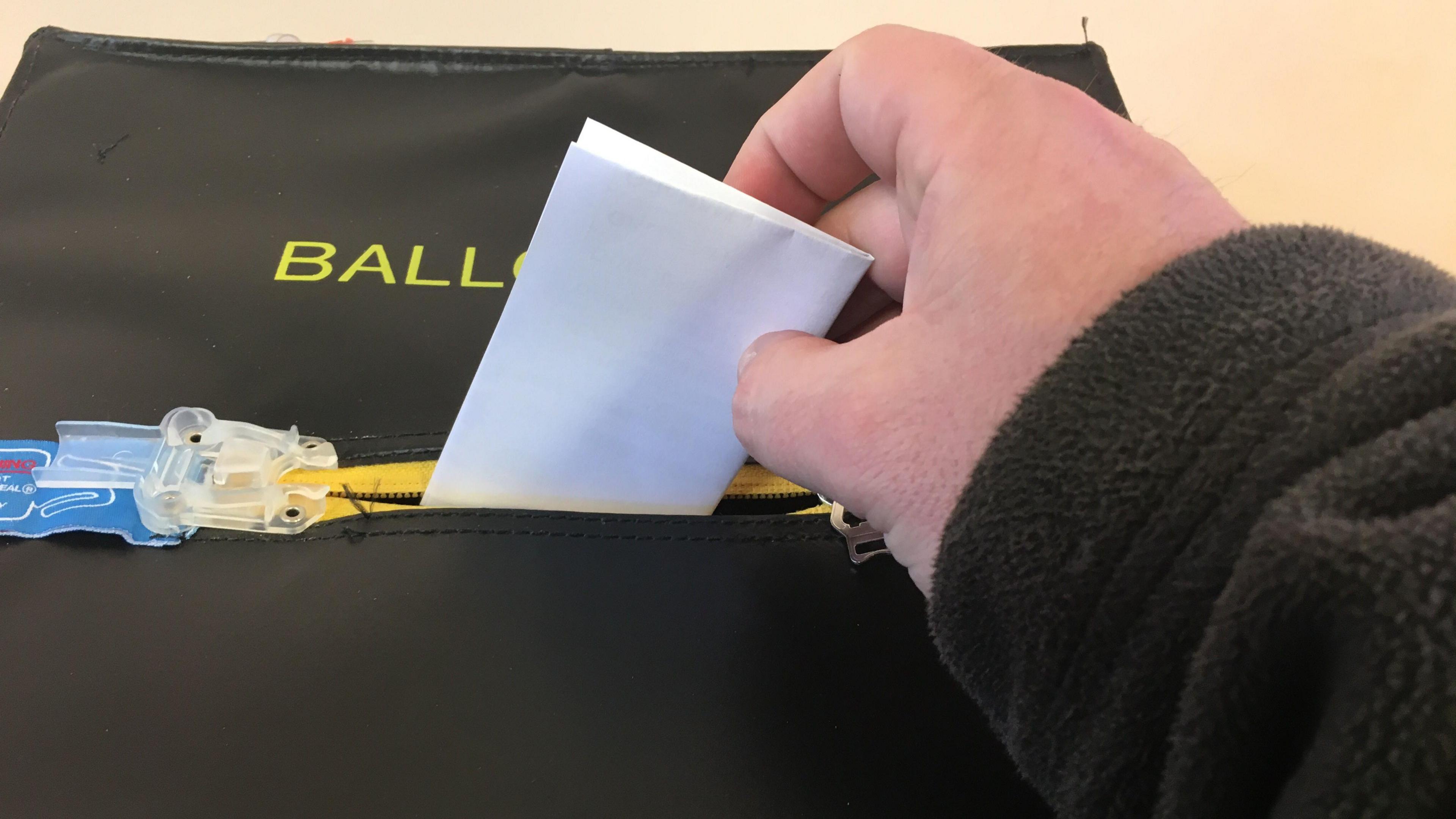 A hand placing a piece of paper into a black box which has the word ballot on it in yellow writing