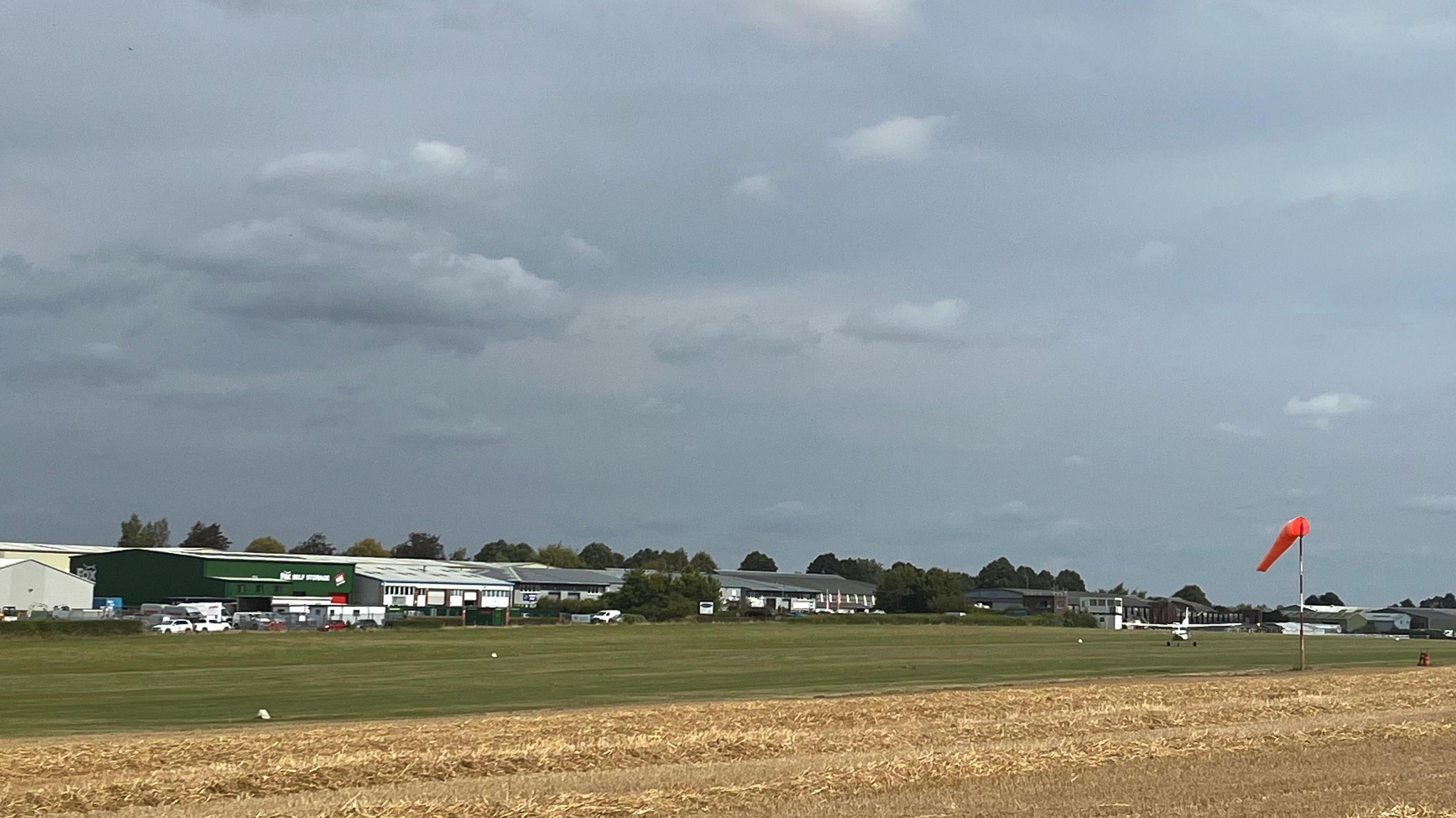 A view of the airfield under a grey sky. A mown yellow crop field with a green airstrip next to it. Aviation buildings in the background. One small plan on the grass airstrip.