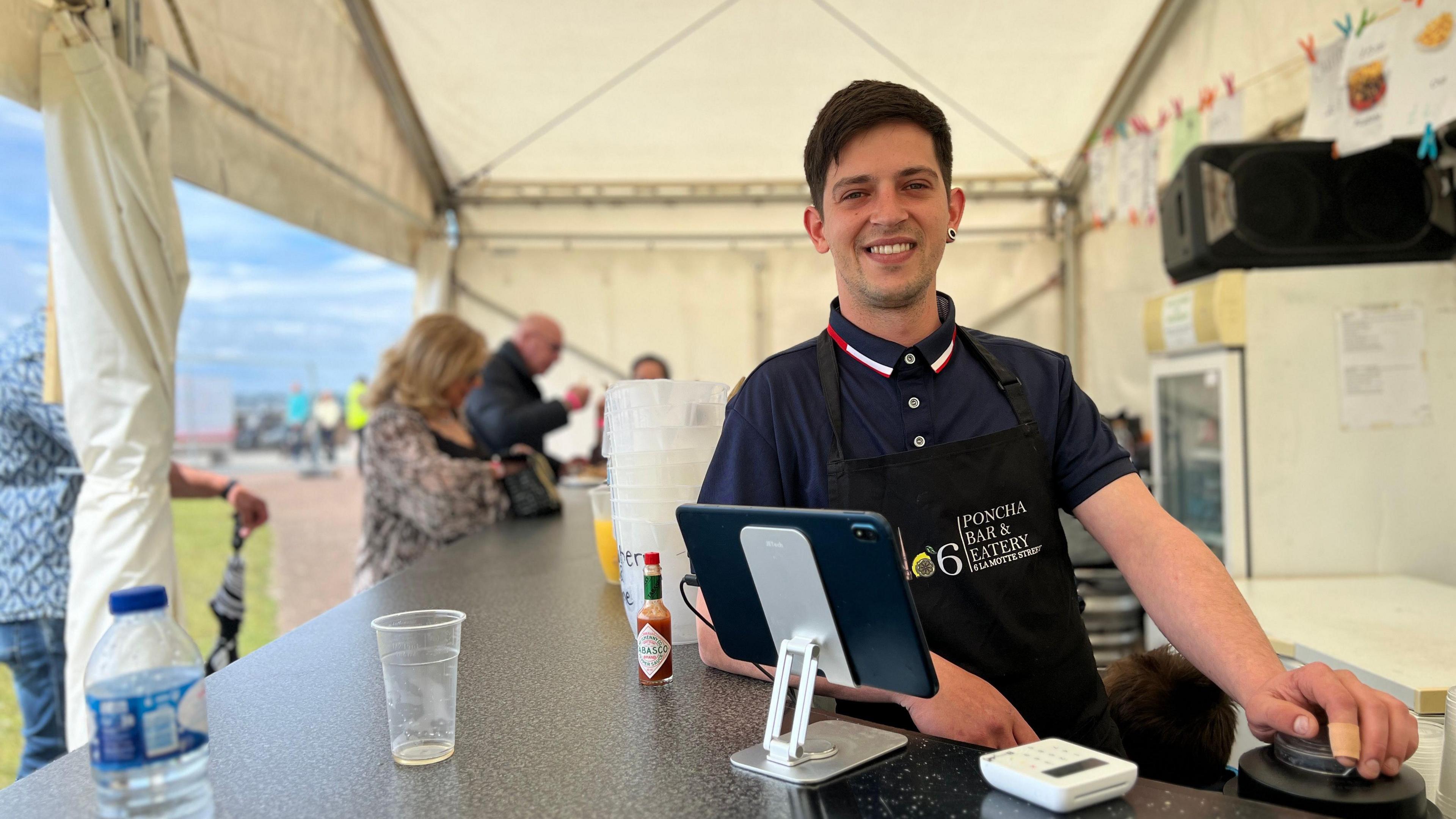 Man smiles at the camera behind a bar while some people enjoy food and drink behind him