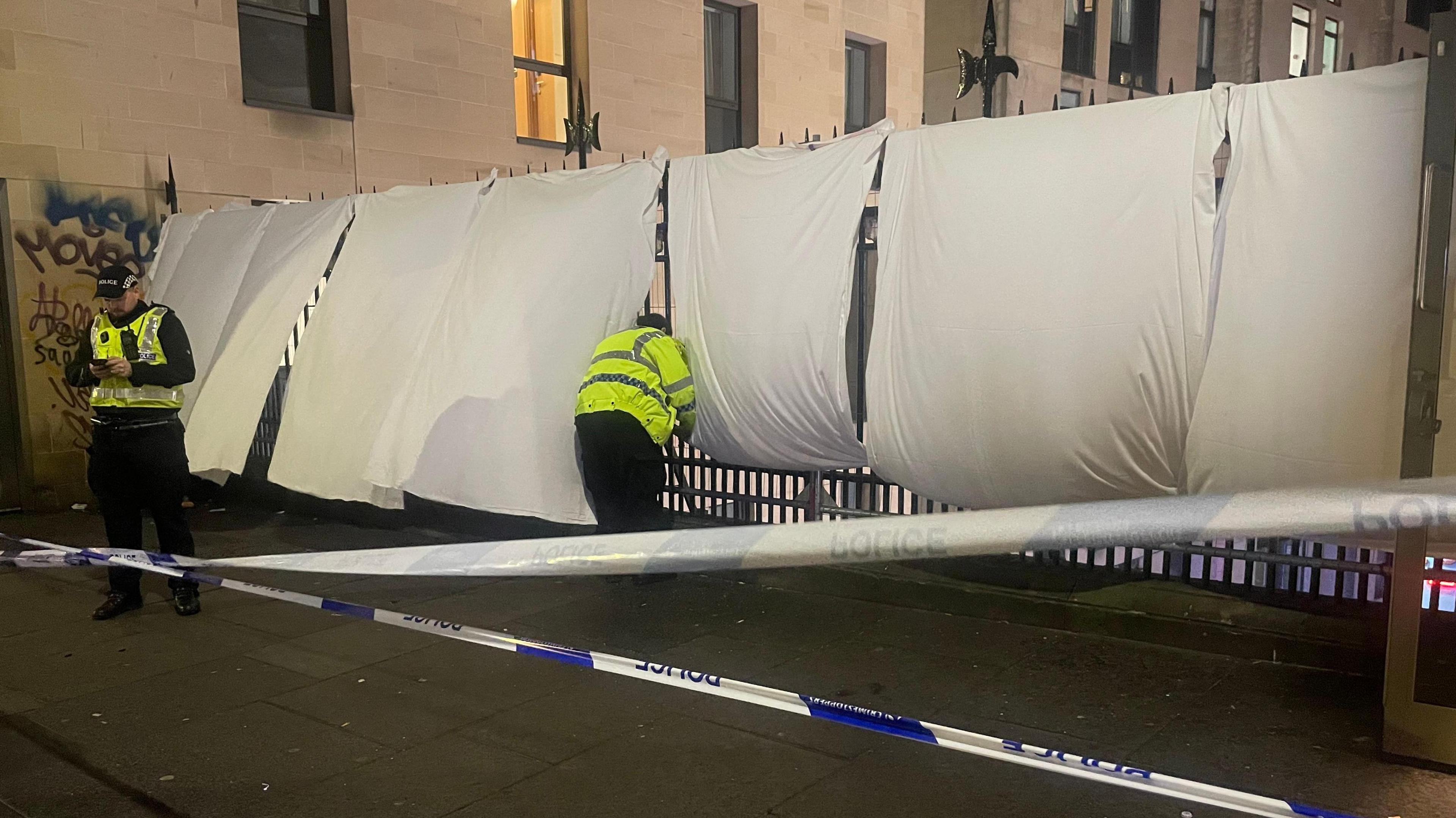 A row of white sheets are erected on the railings of a bridge by yellow jacketed police officers while police tape is visible in the foreground