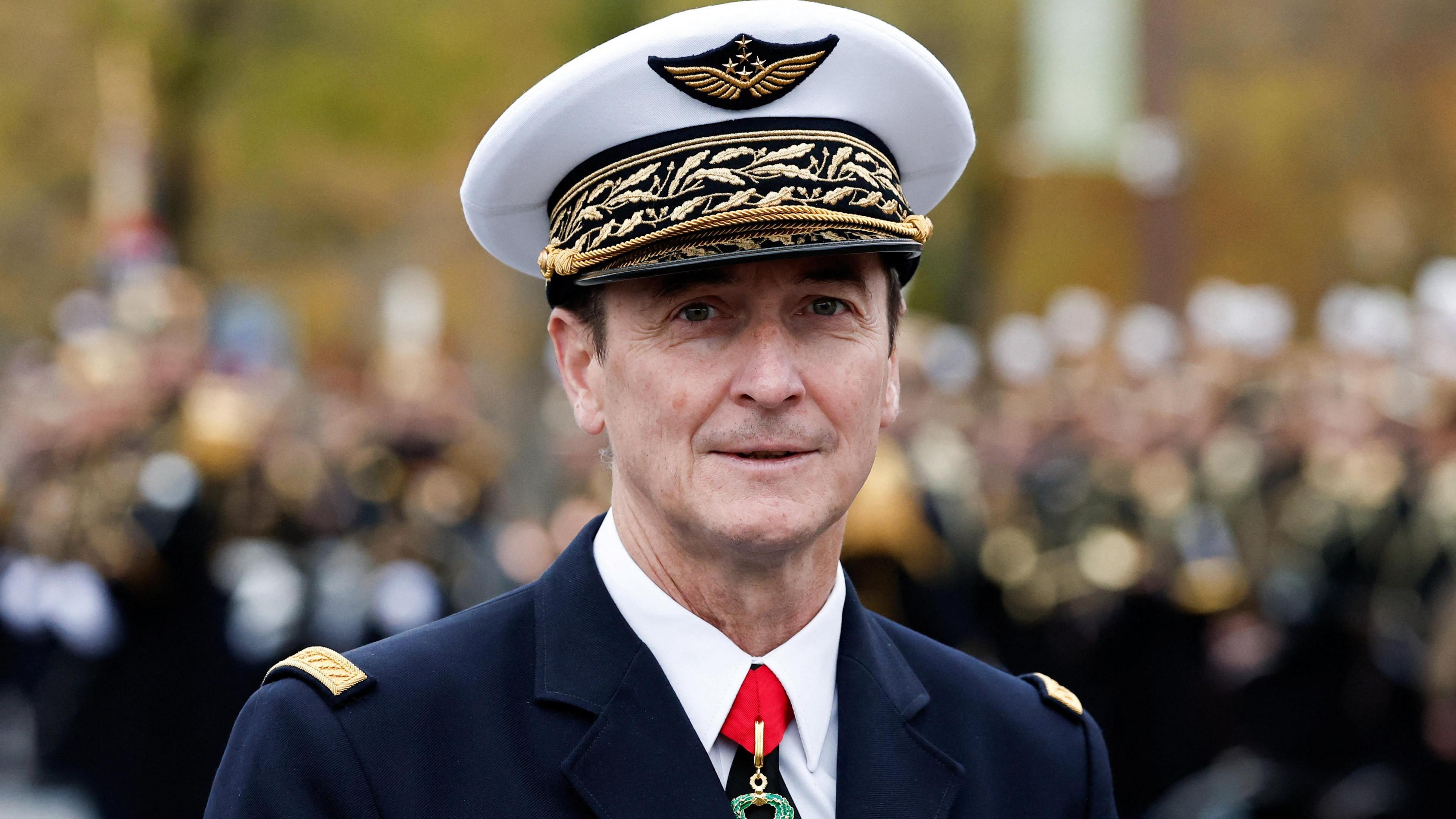 France's Chief of the Defence Staff Fabien Mandon wearing military uniform. He is wearing a white hat with golden winged symbol on it and a black and gold trim. His suit is blue with gold trim on the shoulders 
