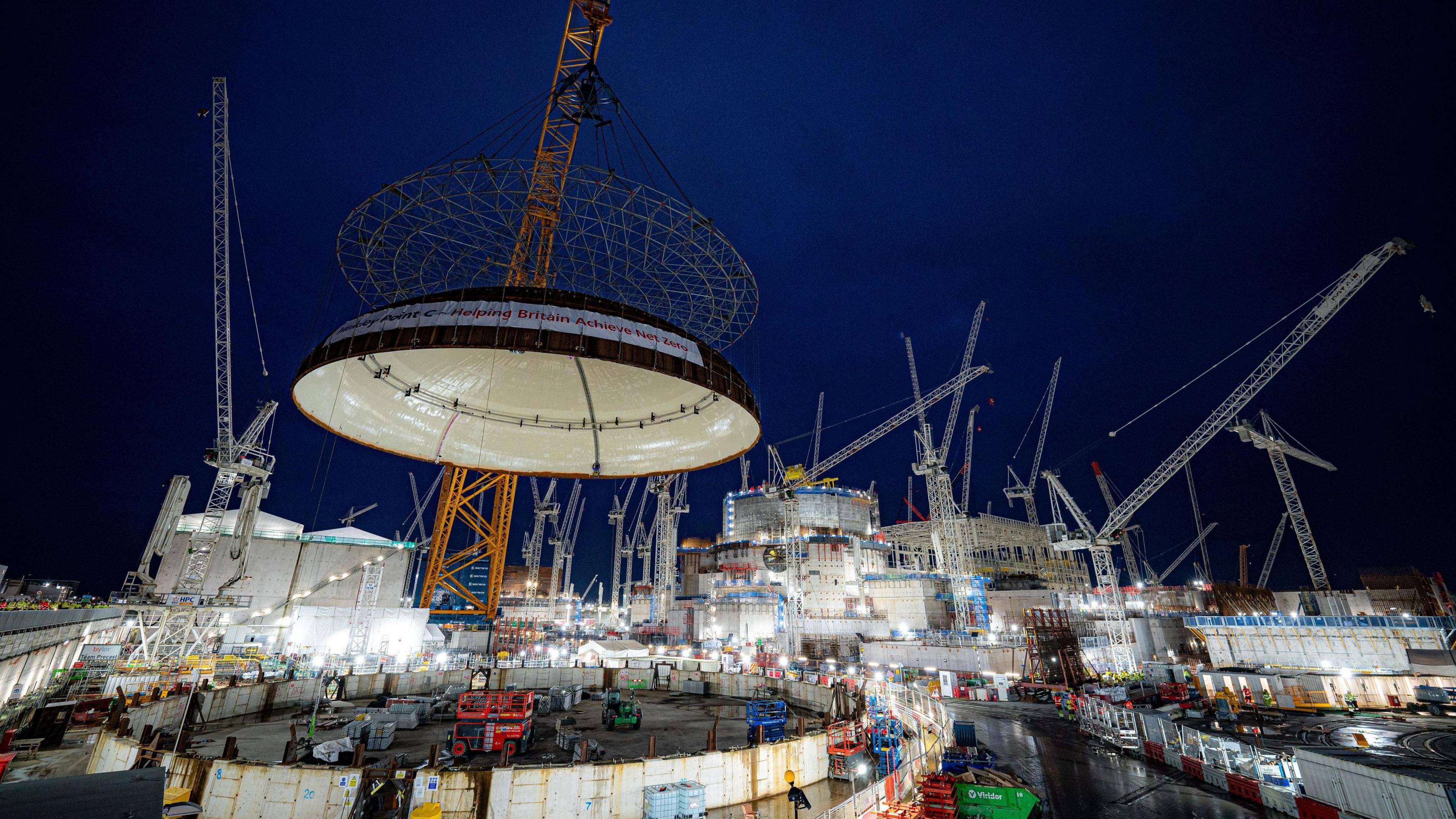 A giant crane lifts the huge dome roof onto the reactor building at Hinkley Point C. It's dark but the forest of cranes is lit up.