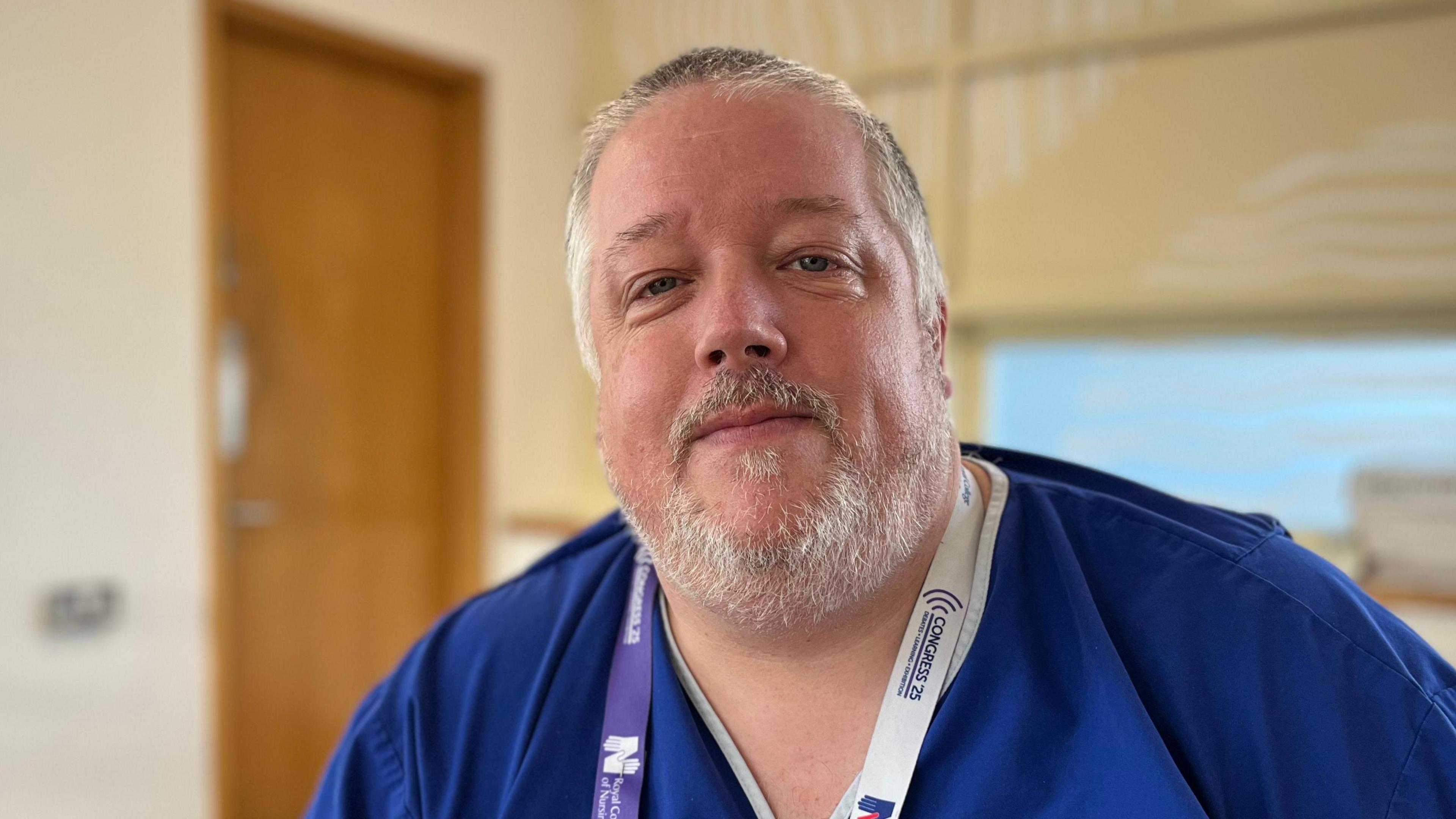 Nick Dove - A man with black and white hair and a black and white beard. He is wearing a Royal College of Nursing lanyard. He is wearing blue scrubs.