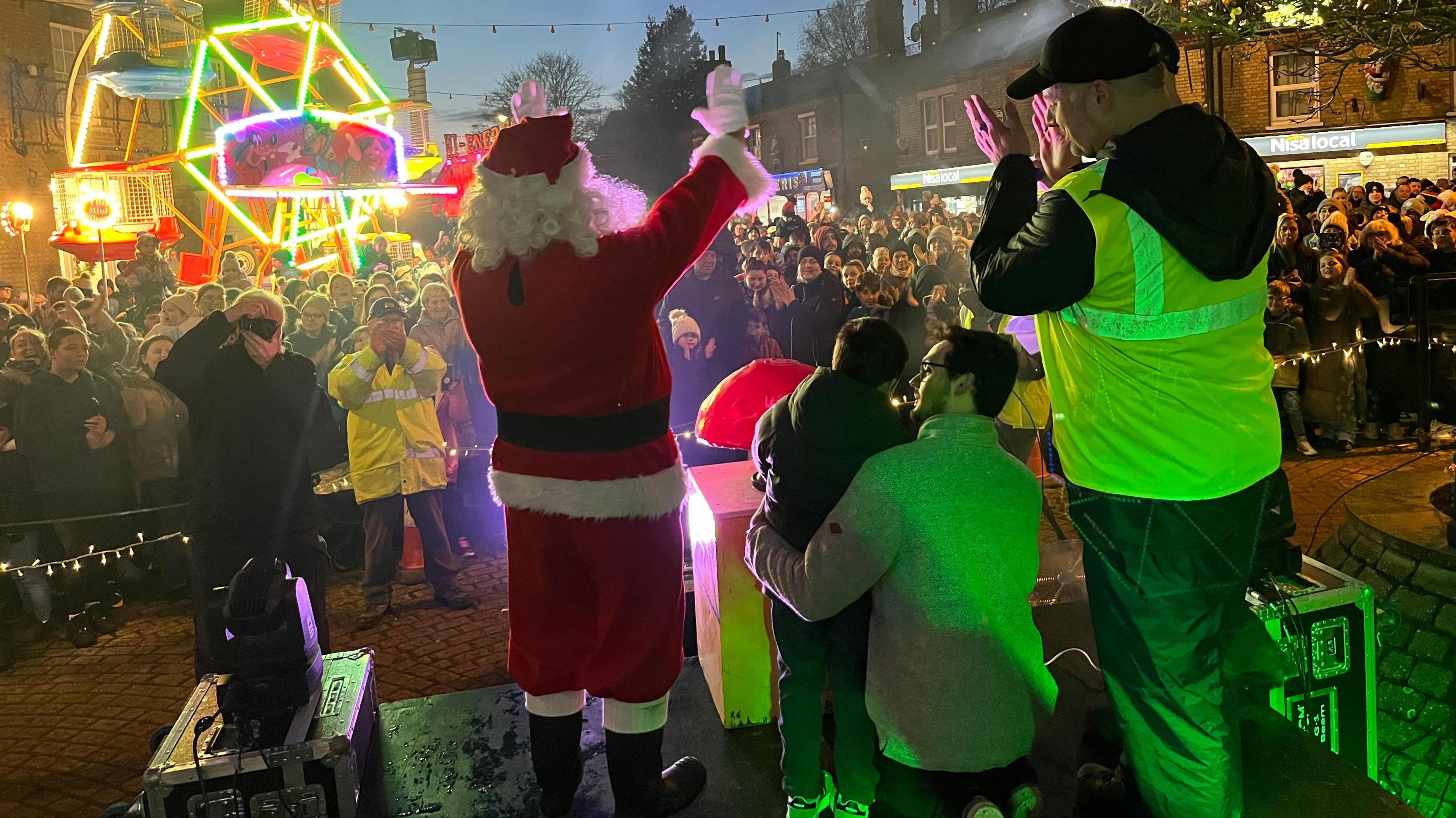 A Christmas lights switch on event, with Santa waving at a large crowd from a stage that also has three other people on it, including a small boy who helped push the red plunger to turn the display on.