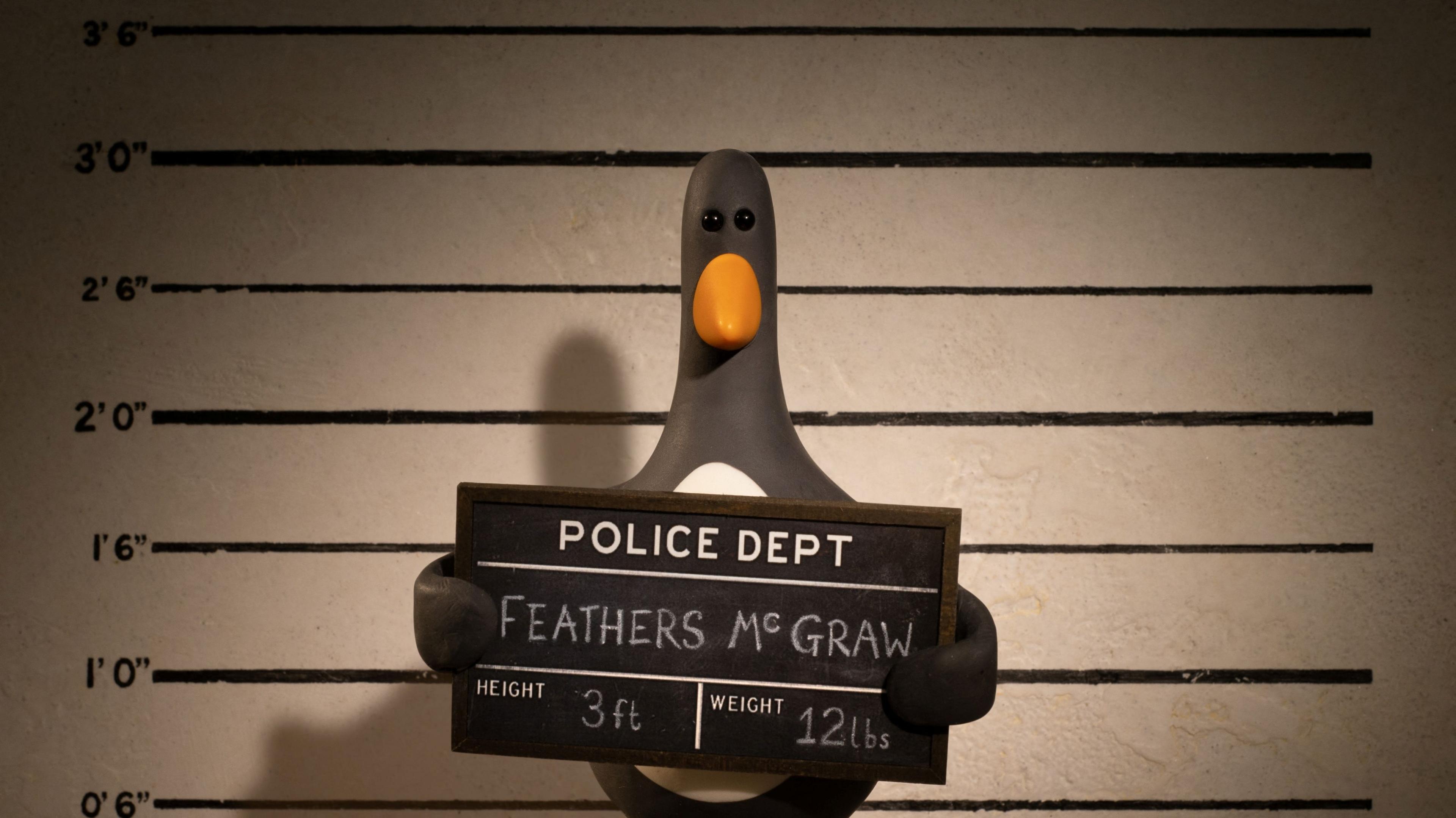 Production still of Feathers McGraw in a police mugshot scene. A penguin stands against a height guide and holds a board which reads "Police Dept: Feathers McGraw". 