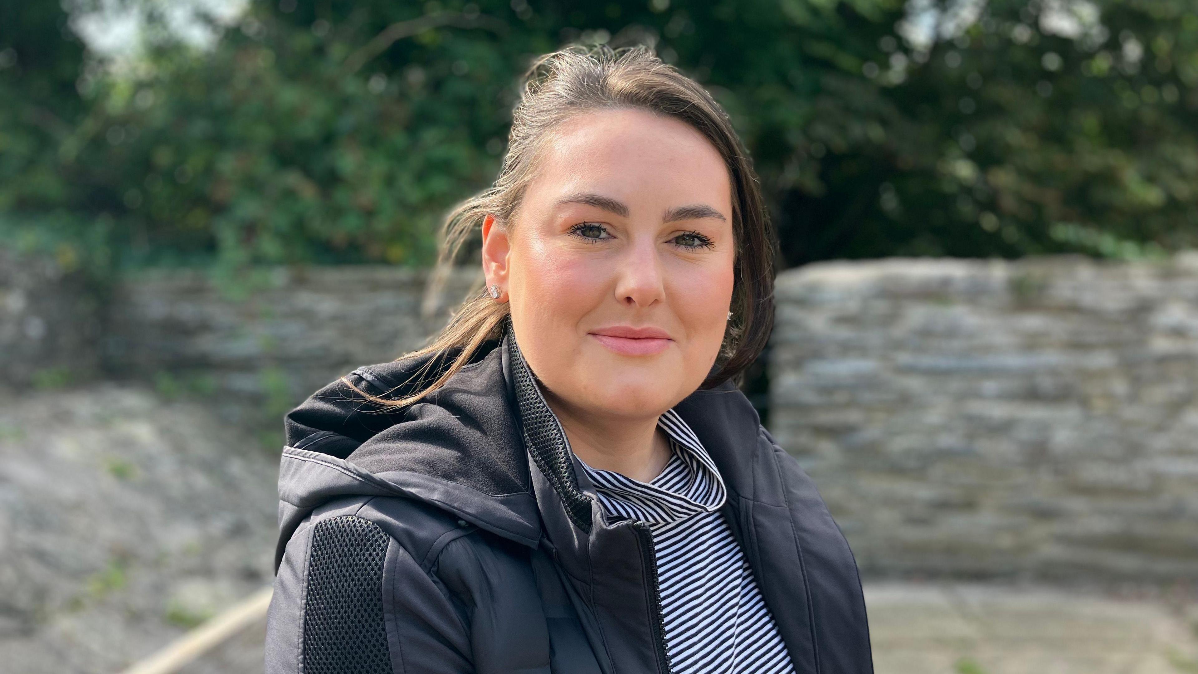 Ffion smiling looking at the camera. She stands in front of a grey wall and trees. Ffion is wearing a navy and white striped top and a black coat. She has medium length dark brown hair which is blowing in the wind. 