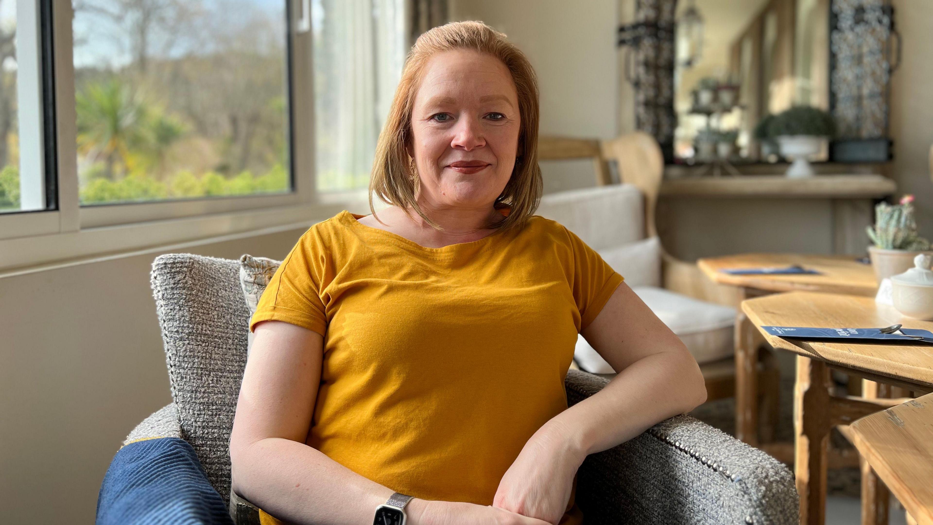 Lorna Pirozzolo is smiling at the camera with her hands folded in front of her. Wearing a bright orange T-shirt she is sitting in an armchair with a table to her left and a mirror behind her. There is window to her right through which a garden can be seen.