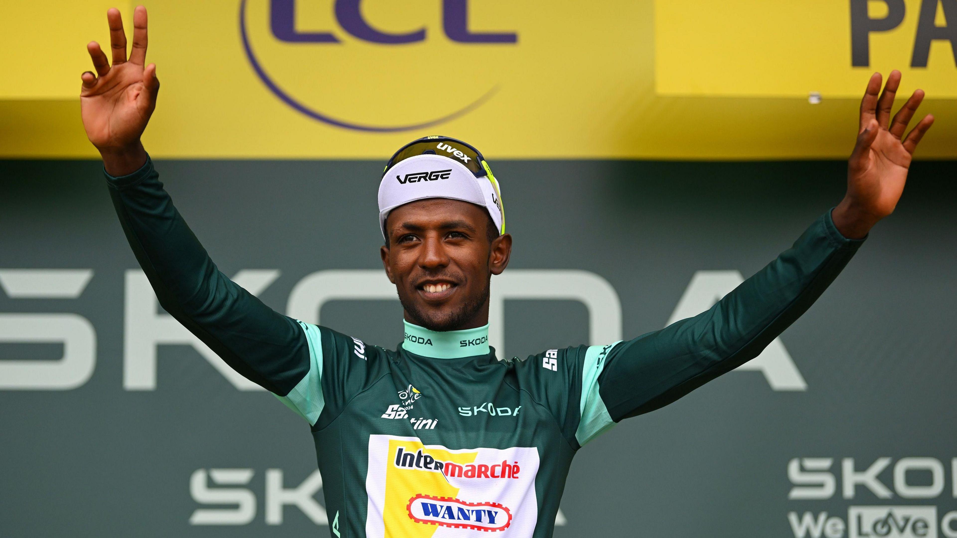 Biniam Girmay, visible from stomach up wearing a long-sleeved green cycling jersey, white cap and sunglasses pushed up on to the top of his head, holds both arms up as he smiles in celebration. He is on a post-race podium, discernible due to the sponsors logos visible on the yellow and green backdrop