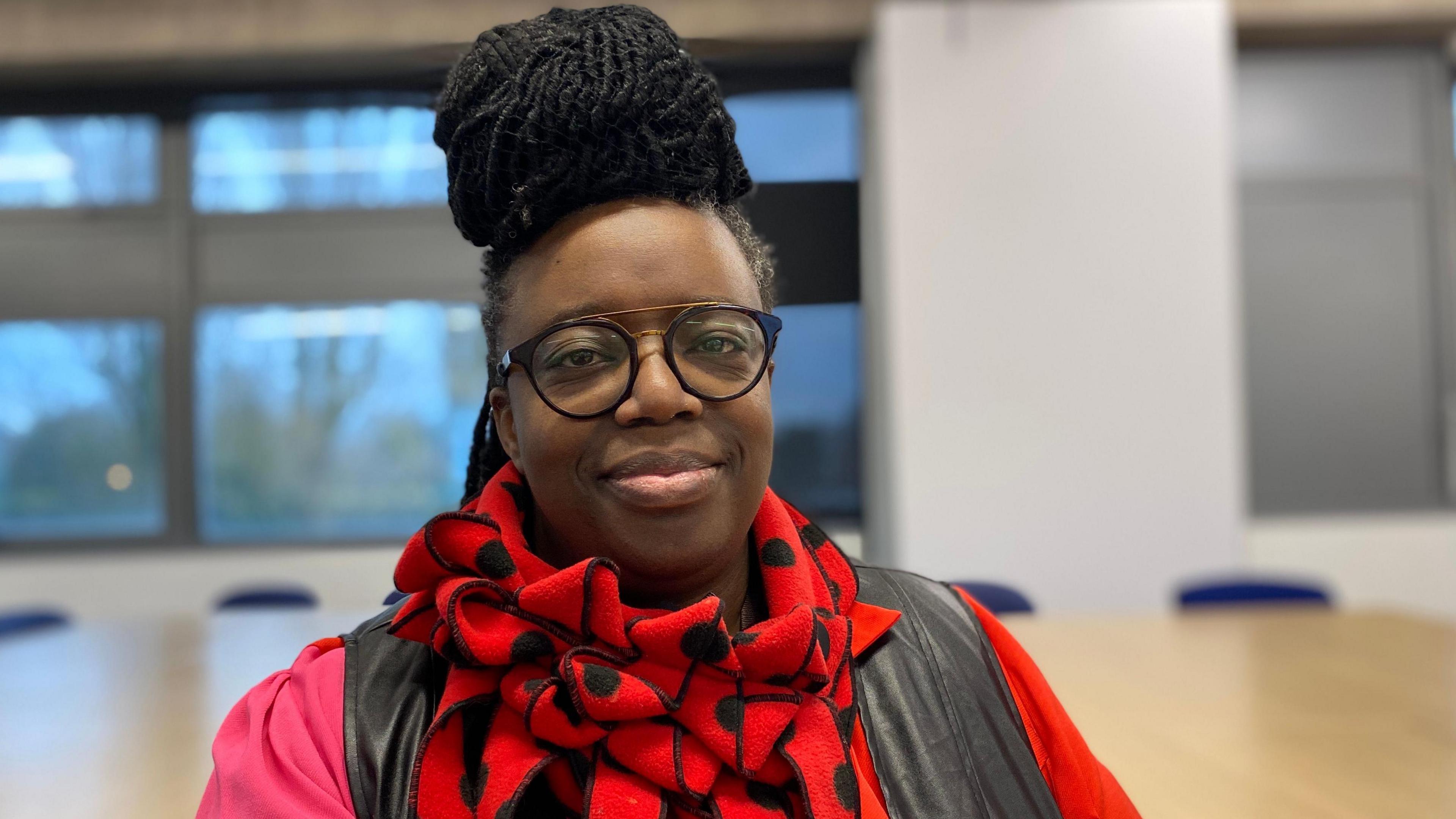 A woman wearing a red scarf with black polka dots and a red top. She has black glasses and dark hair in a bun.