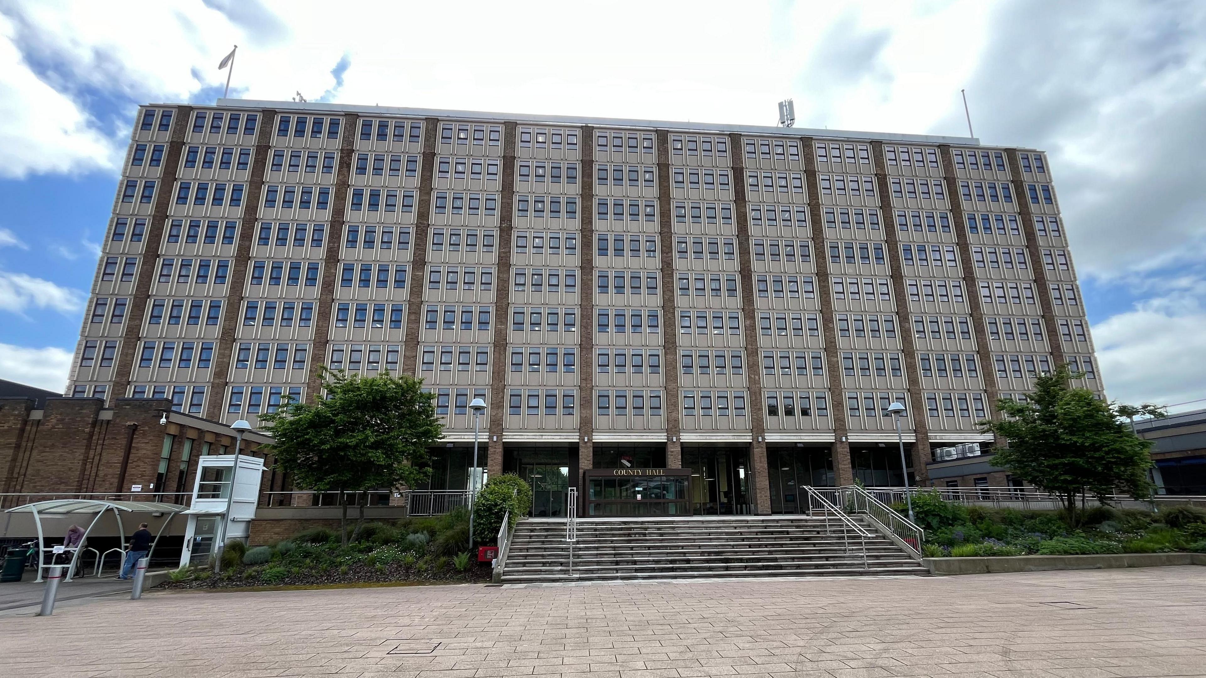 Norfolk County Hall, a tall, modern grey  concrete buildings with many rows of windows, wide steps going up to it from a paved court yard and two trees in leaf on either side of the steps