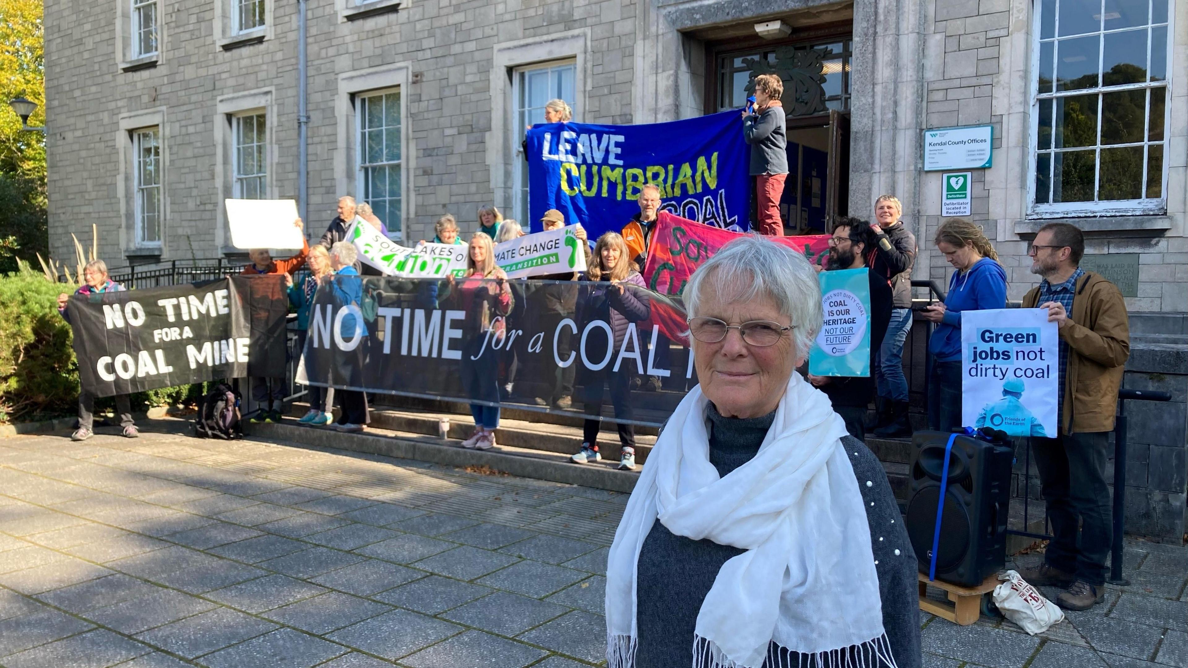 Maggie Mason, who has short, white hair and glasses, stands in front of campaigners who are holding signs and banners protesting against the Whitehaven coal mine proposal.