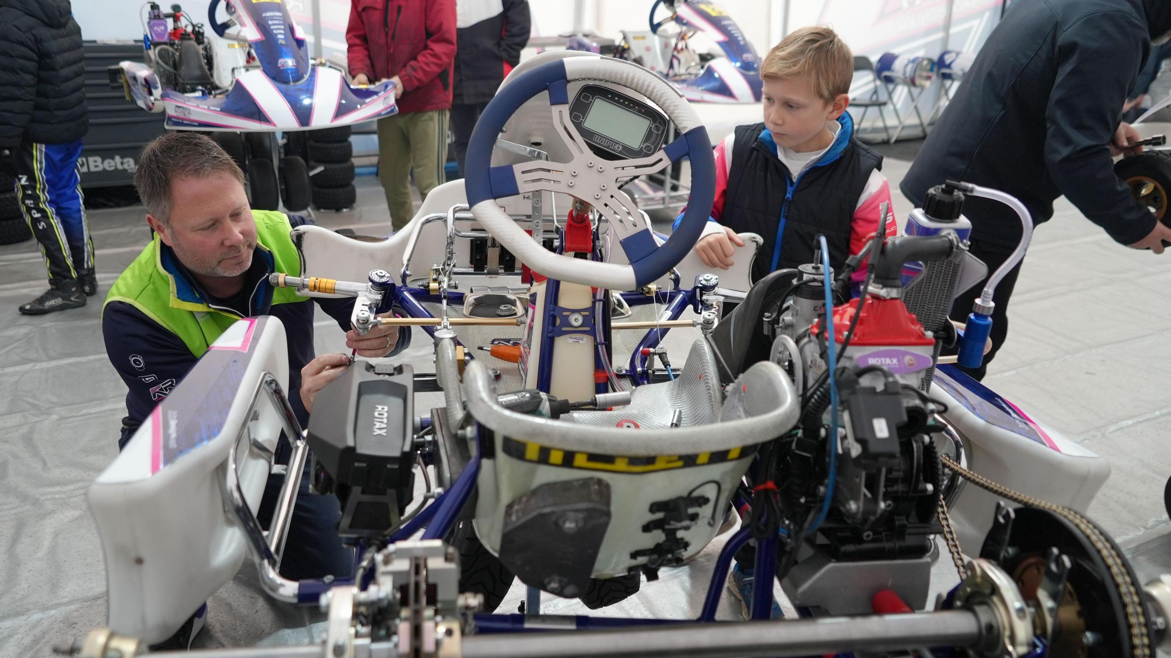 Josh watches as his father, wearing a hi-vis top, makes adjustments to his kart. Other people can be seen working on karts behind them.