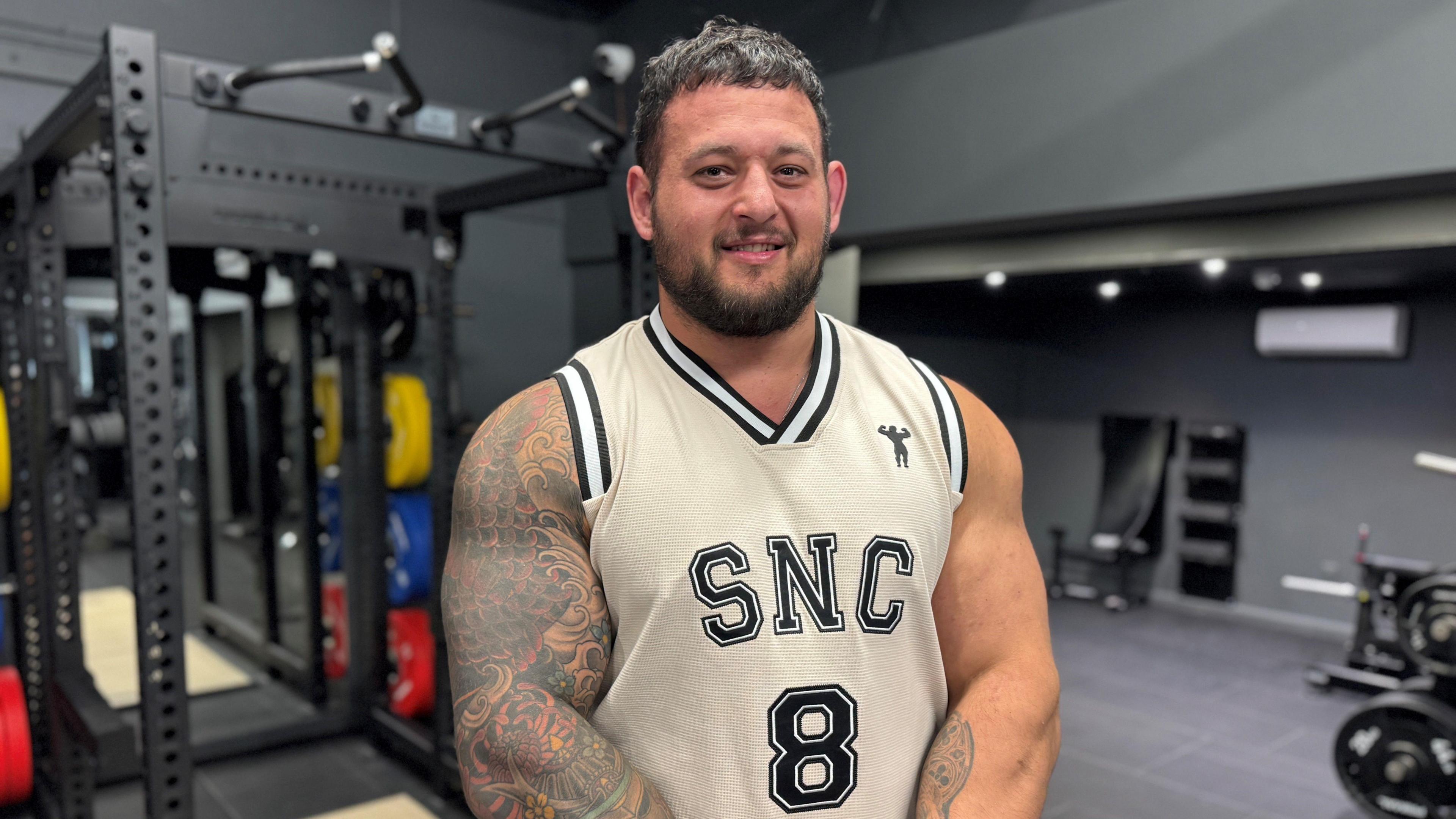 Shane Flowers stood in a gym, wearing a white vest top and sat in front of some weights