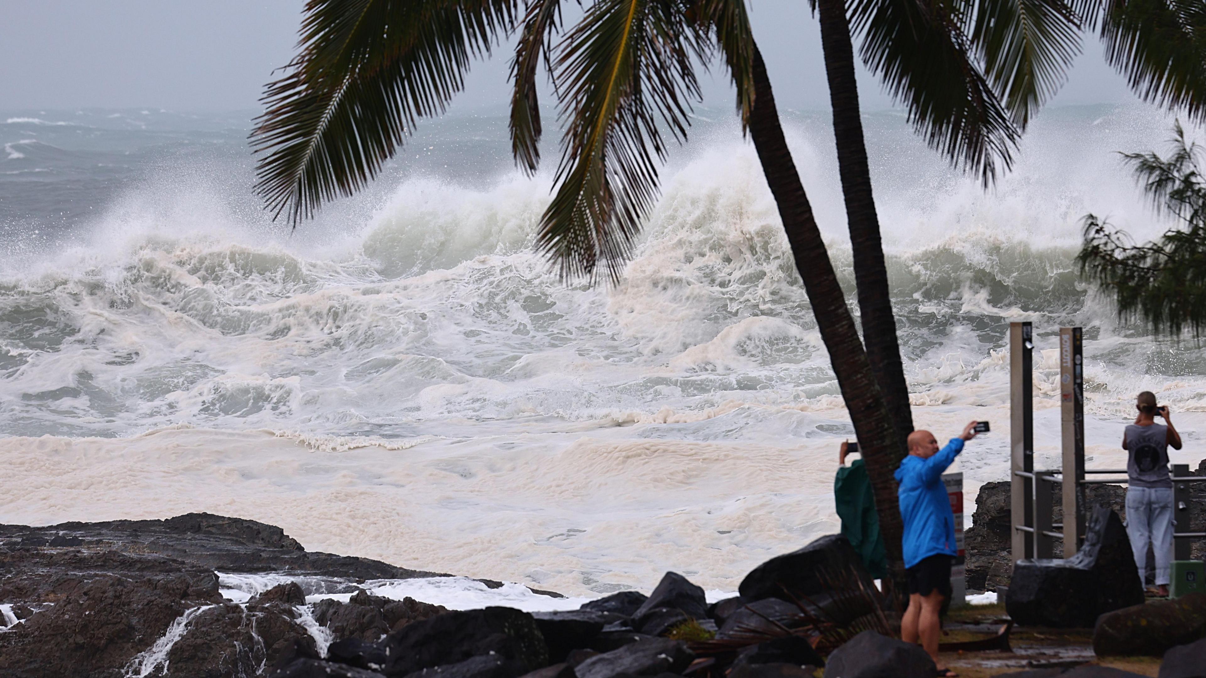 People are seen at Snapper Rocks on the Gold Coast, Queensland, Australia. Three people take photos with their phones by a palm trees in front of high waves in the sea. 