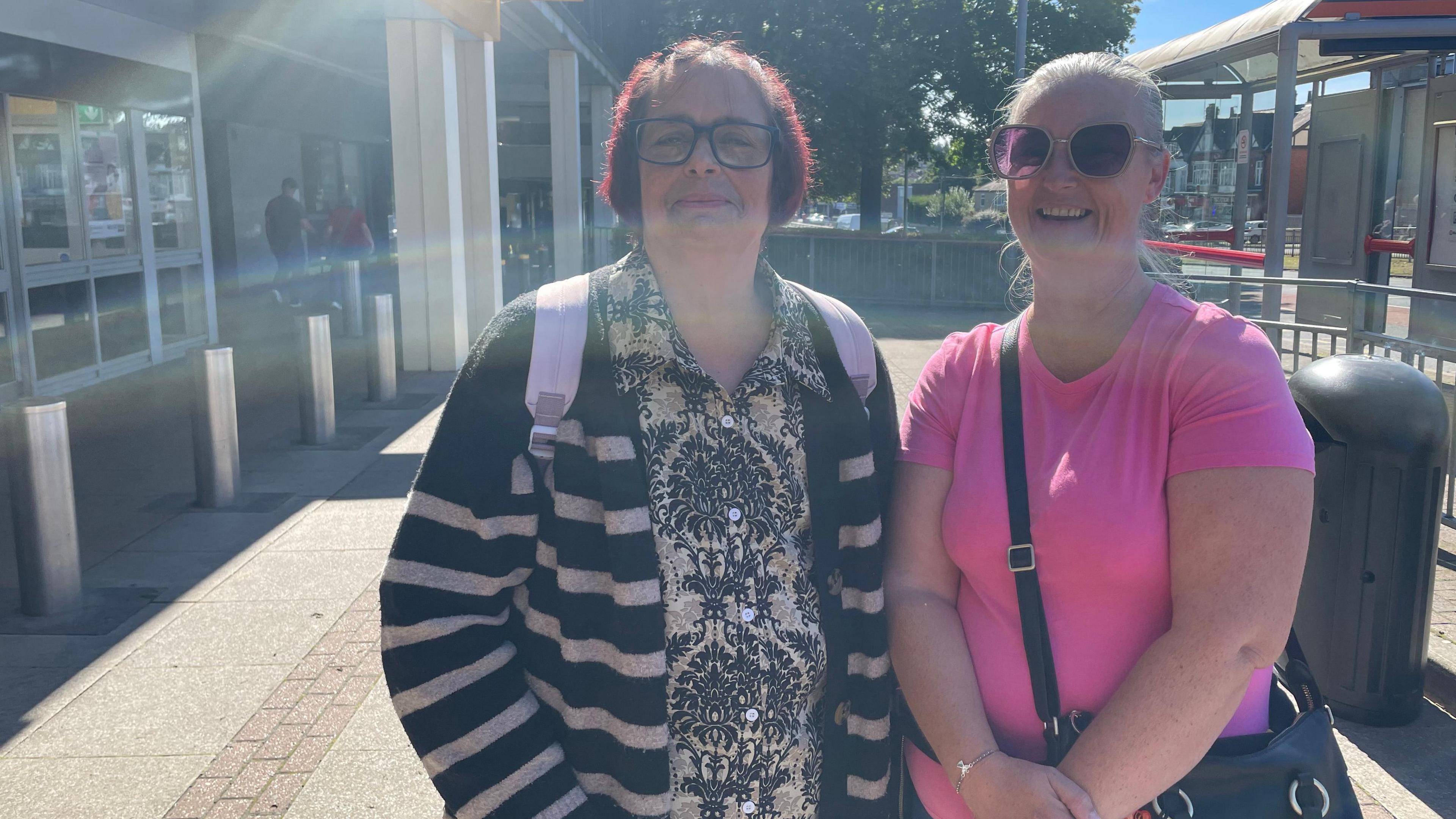 Jean Barnbrook and Emma Bland standing in bright sunlight near a bus stop and bollards