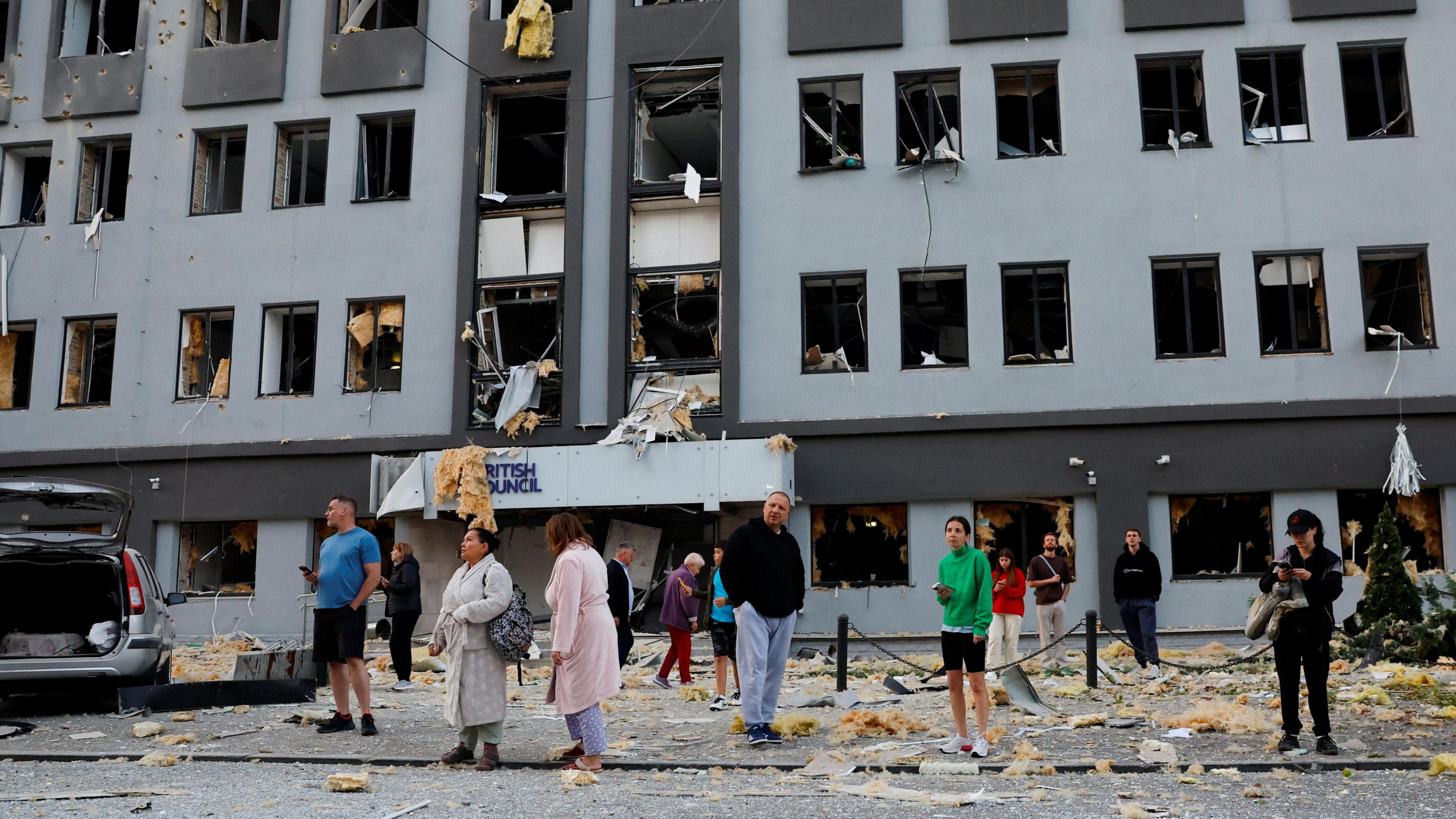 People in pajamas and dressing gowns stand outside a multi-storey building which was hit by a Russian missile and drone strike. All of the windows have been blown out, and the pavement is covered in bits of rubble and insulation. 