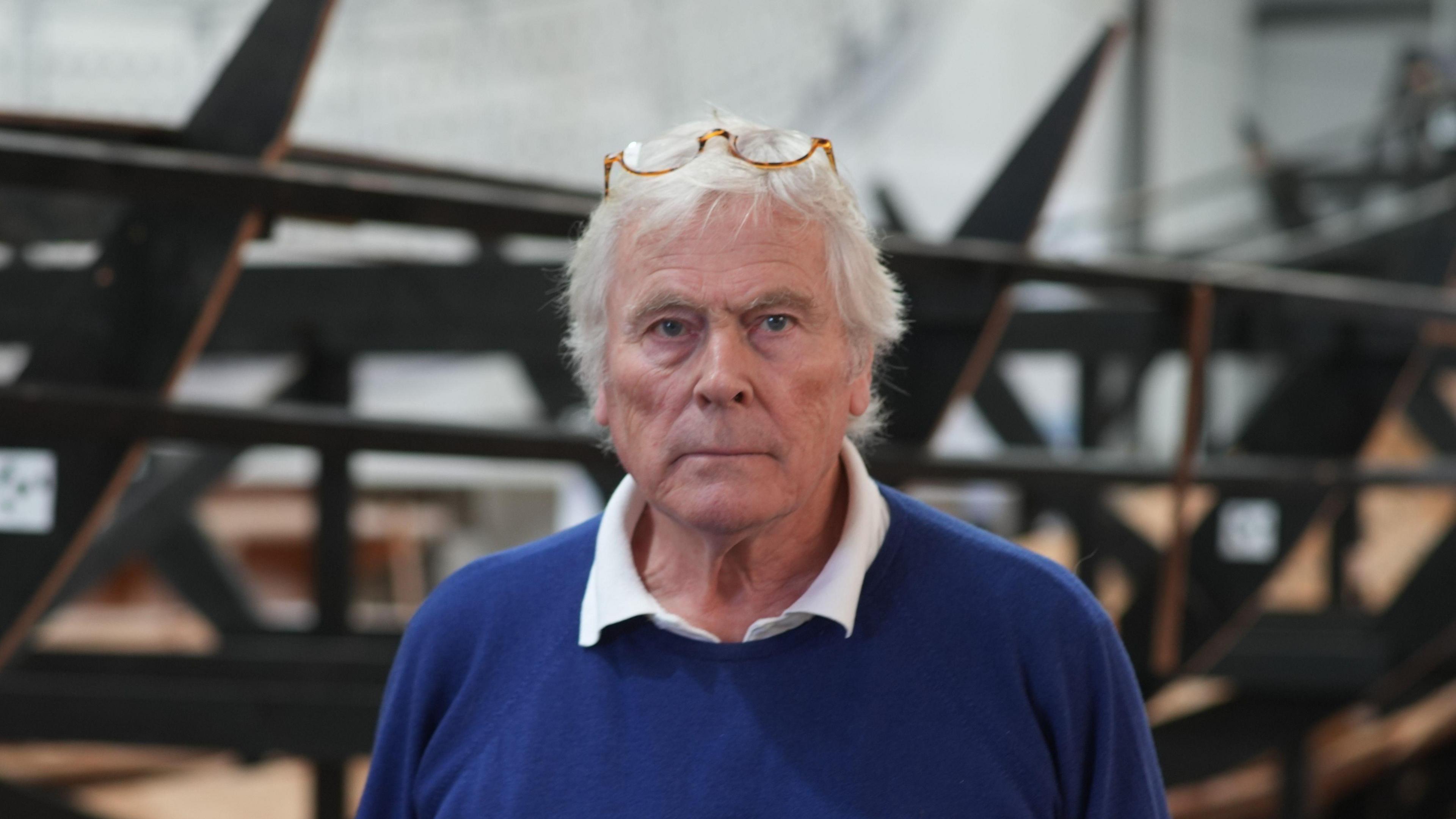 Sean McMillan, trustee and chair of SHSC, is pictured facing the camera. He is slightly smiling while standing in front of the ship replica. He has glasses resting on his head with a blue jumper and white shirt on.