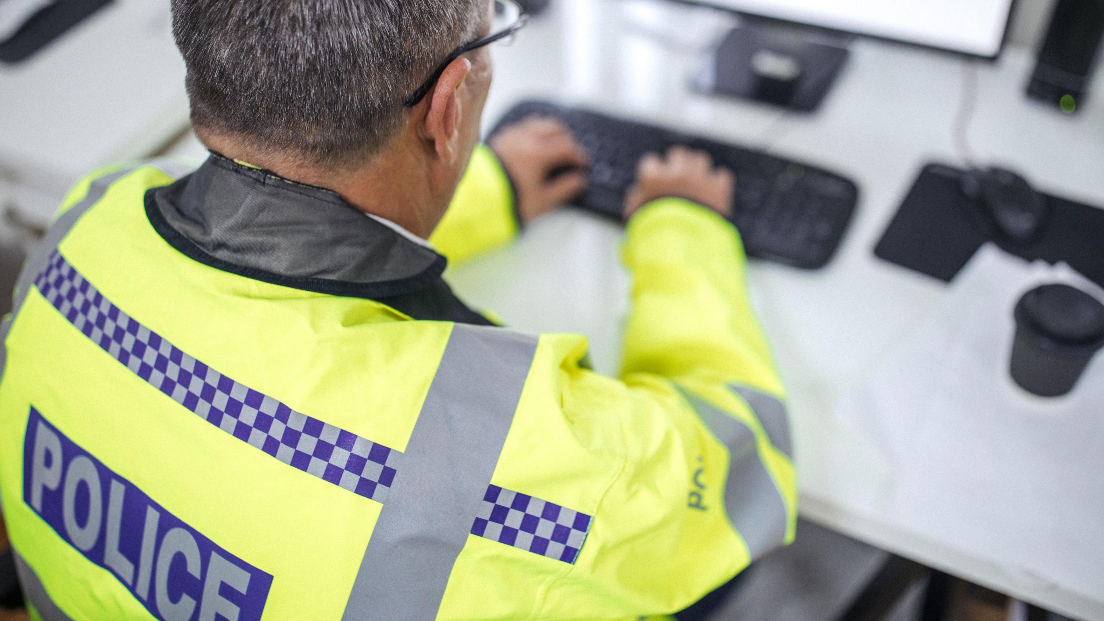 An anonymous Met Police officer wearing a hi-viz jacket with the wording 'police' sat at a desk typing.