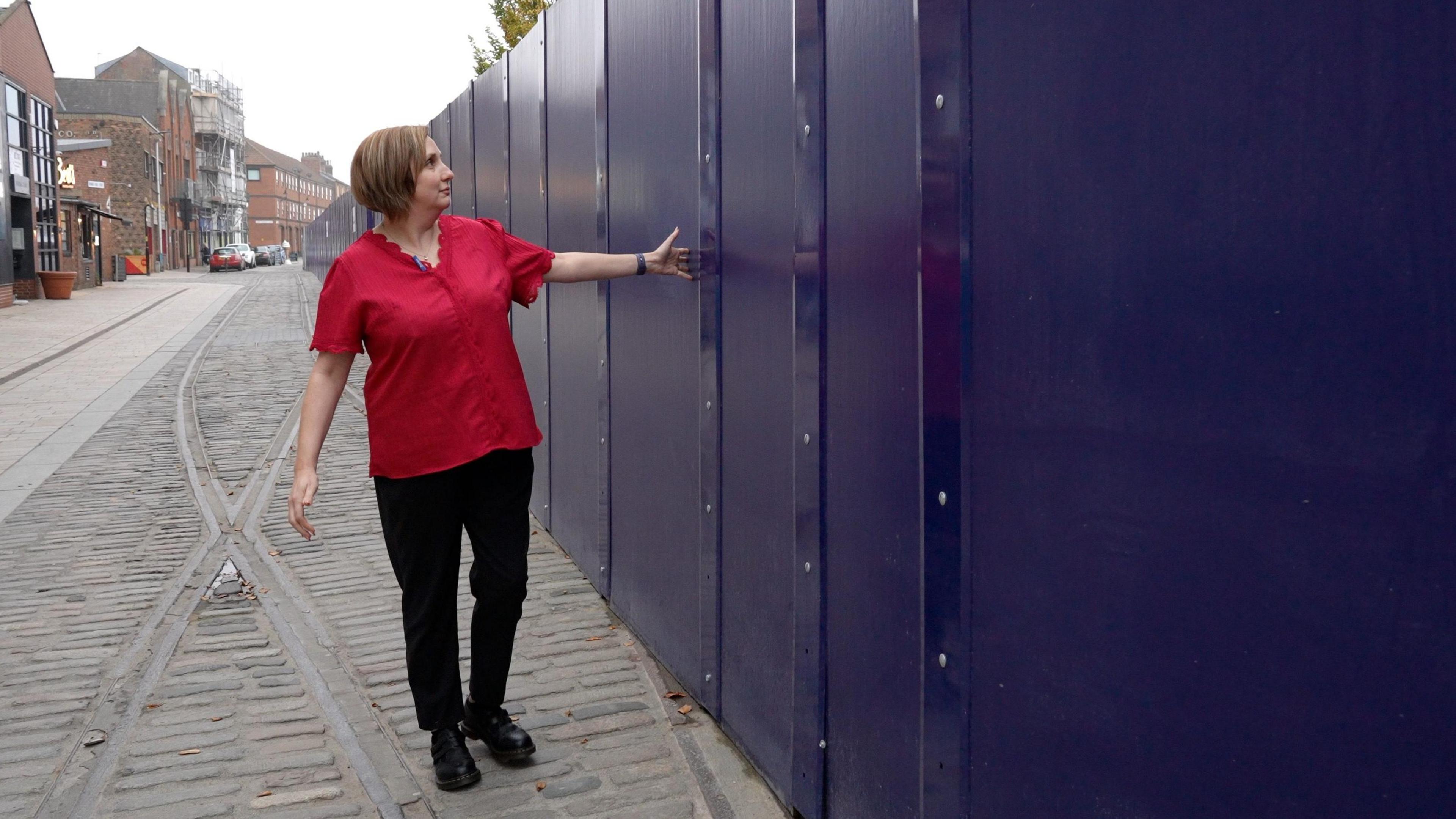 A woman wearing a bright red short-sleeved shirt and black trousers is standing on a cobblestone street next to a tall, solid barrier made of dark blue panels. She is gesturing toward the barrier with one arm extended, as if indicating its length or height. The street has old metal tram lines running along its surface, and older brick buildings are visible in the background on the left side.
