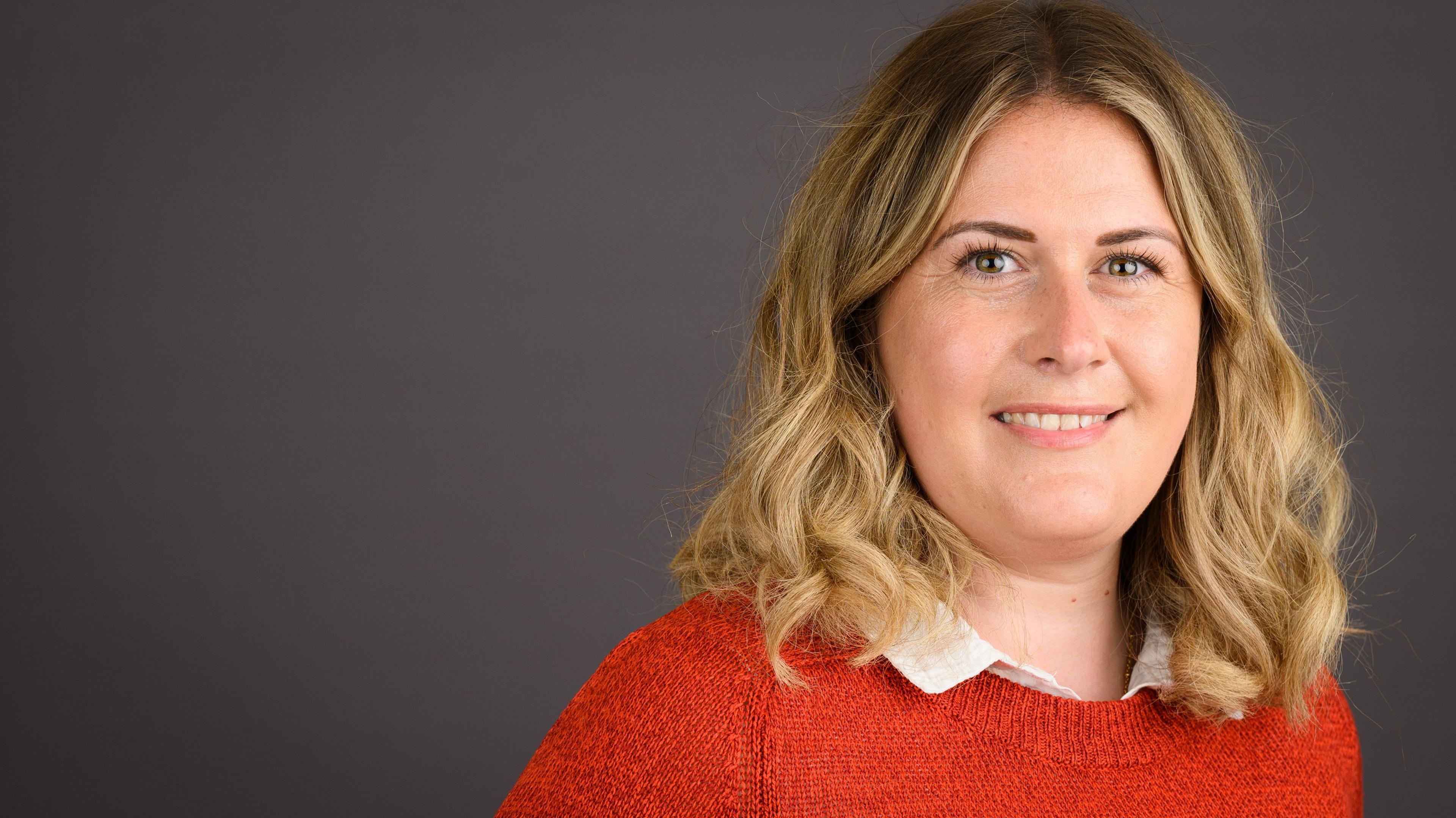 Millie Earl in an orange jumper and in front of a neutral background. Her hair is should length, blonde and wavy