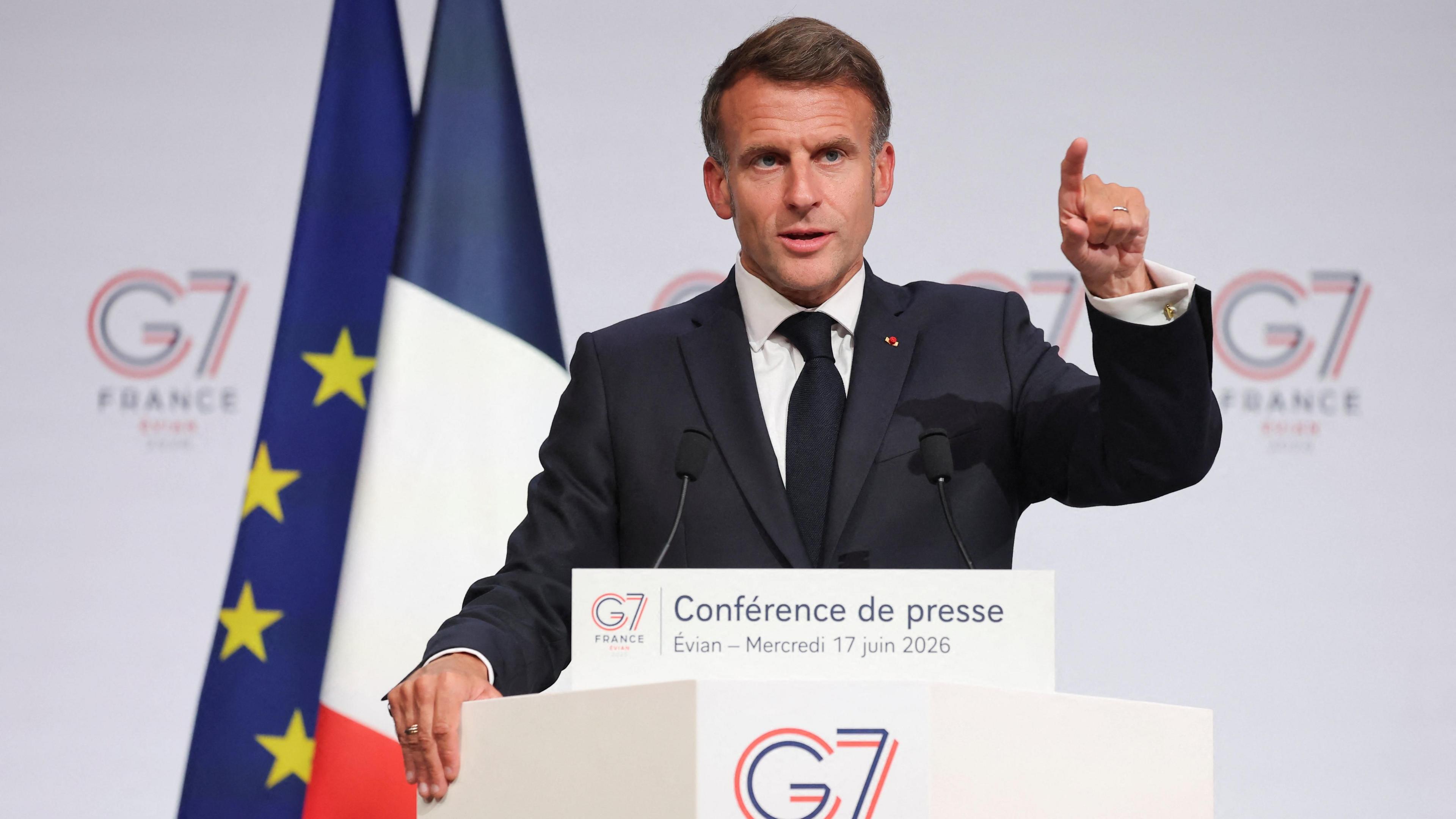 French President Emmanuel Macron holds a press conference during the G7 Summit, he speaks at a lectern and points with one hand.