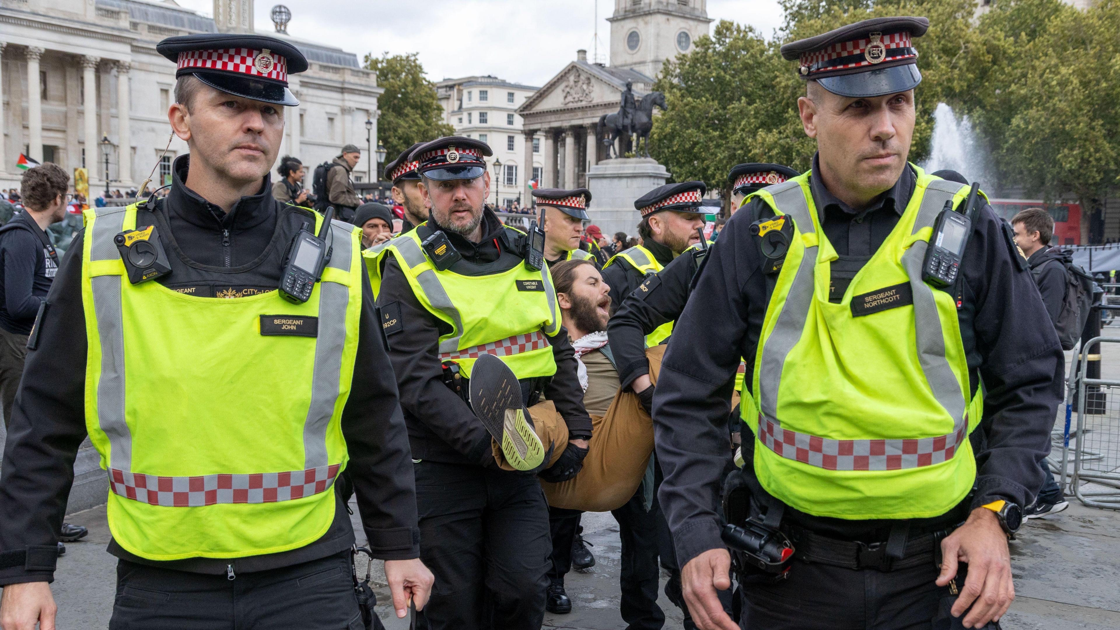 Police officers in high-vis vests arrest a man at a protest at Trafalgar Square and carry him out.