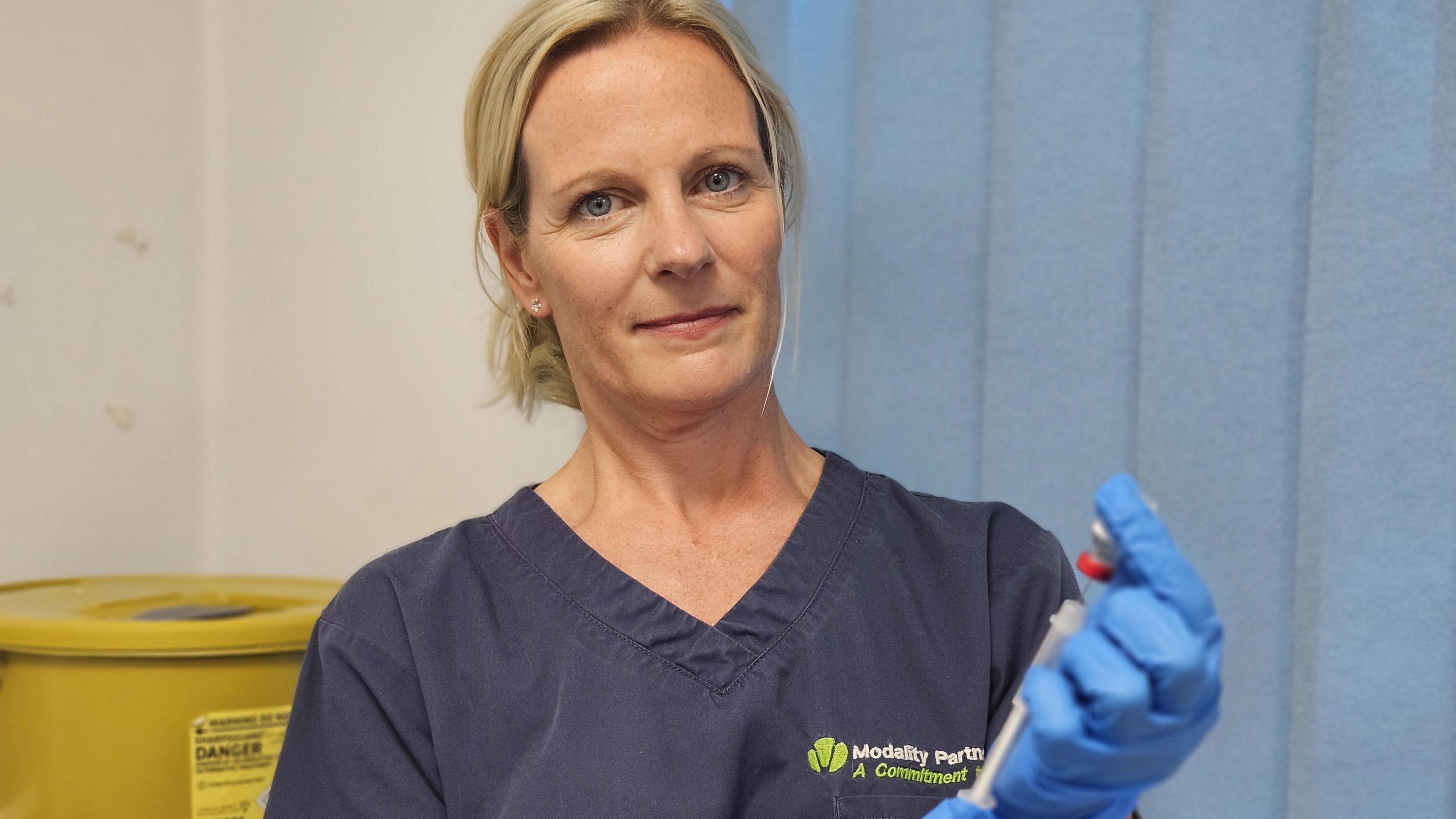 A woman wearing blue medical scrubs and blue gloves poses holding a vaccination needle as she draws from a vial