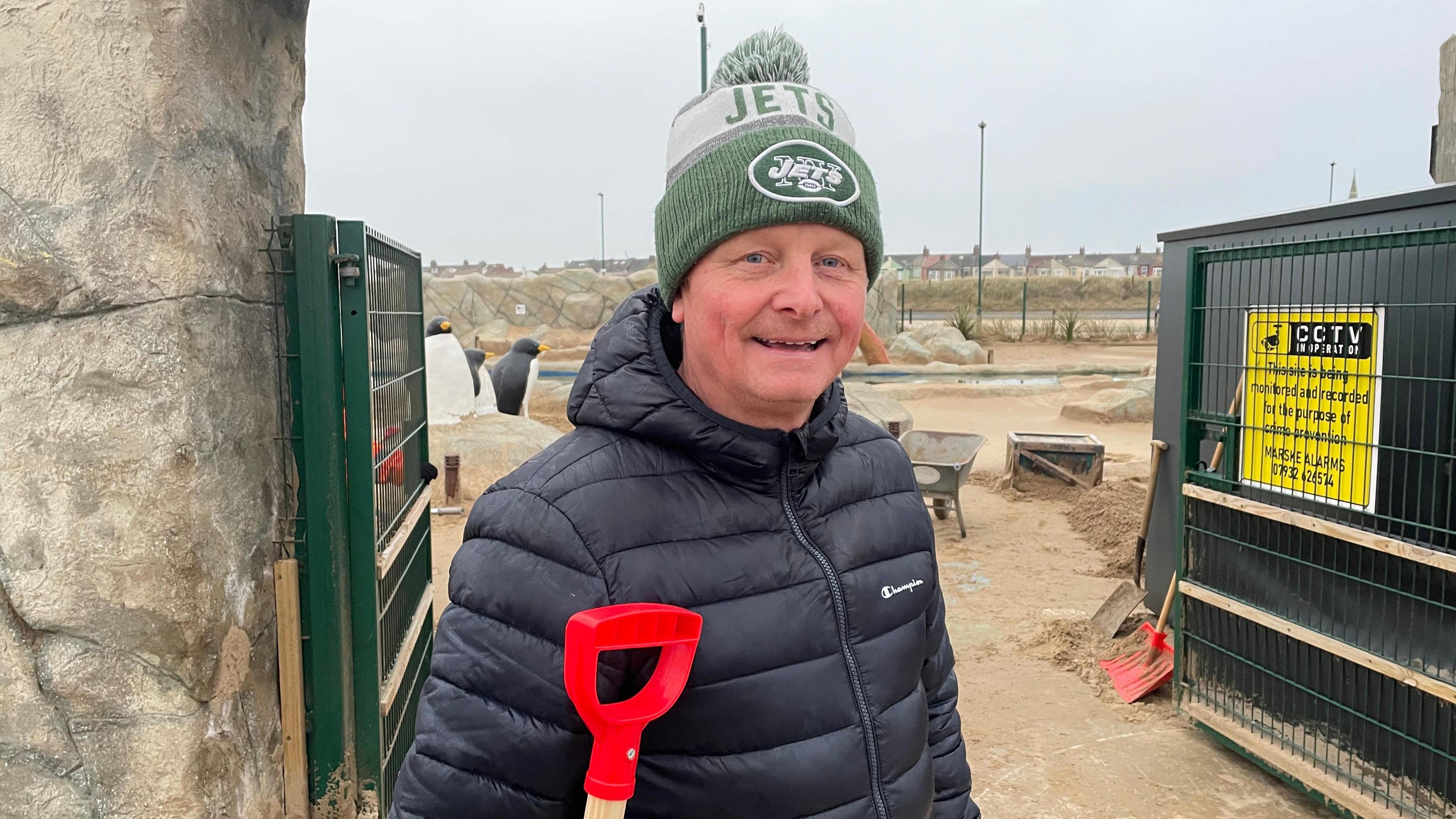 Andrew Smith is standing at the gates if the golf course and is smiling at the camera. He is wearing a big coat and a green woolly hat. He is also holding a red-handled shovel.