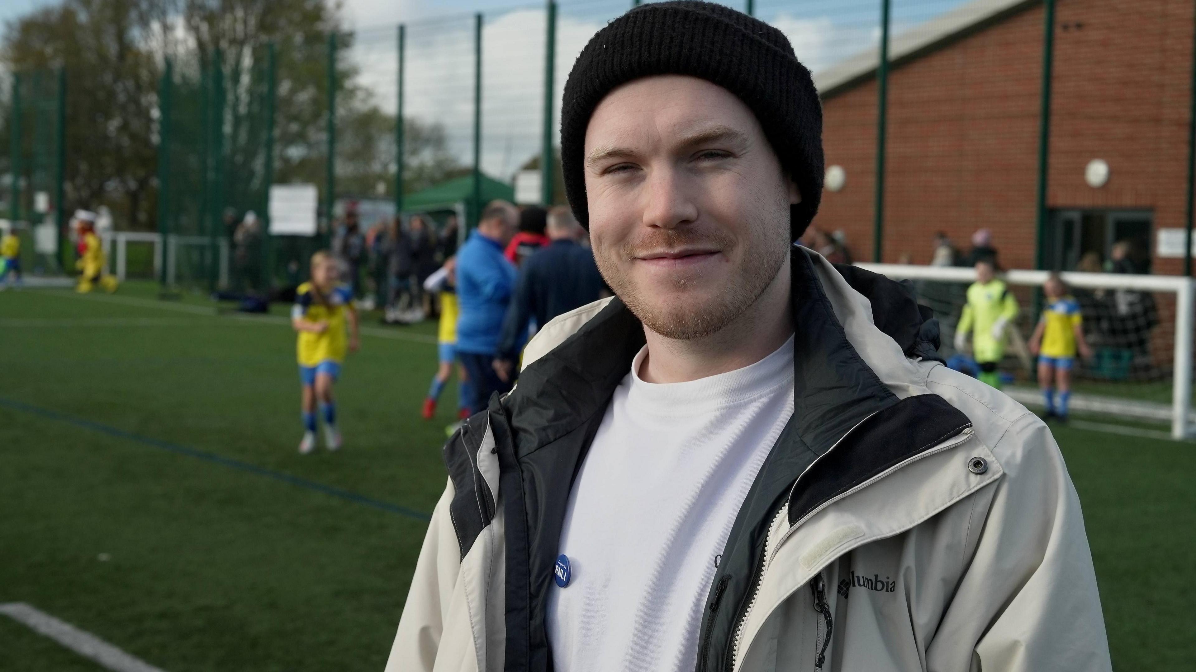 Jamie Scott wears a black beanie and a cream-coloured raincoat. He stands on an artificial turf pitch at Whitley Bay Sporting Club.