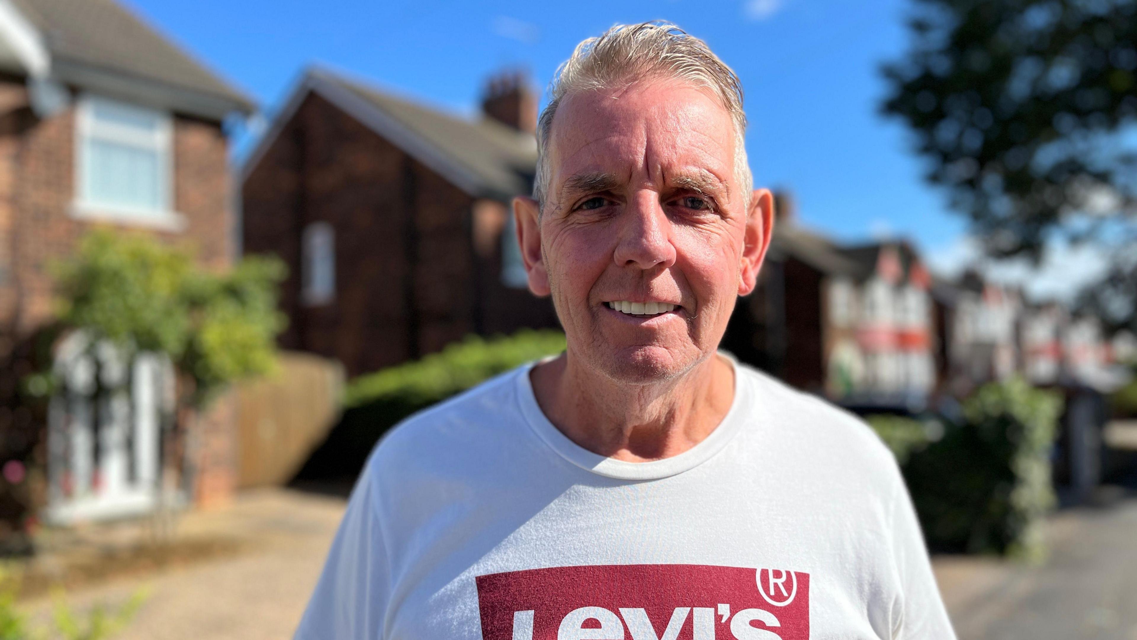 A man with grey hair is wearing a white top with a red and white Levi's logo and looking at the camera smiling. The background is blurred and there is a road with houses in sight.
