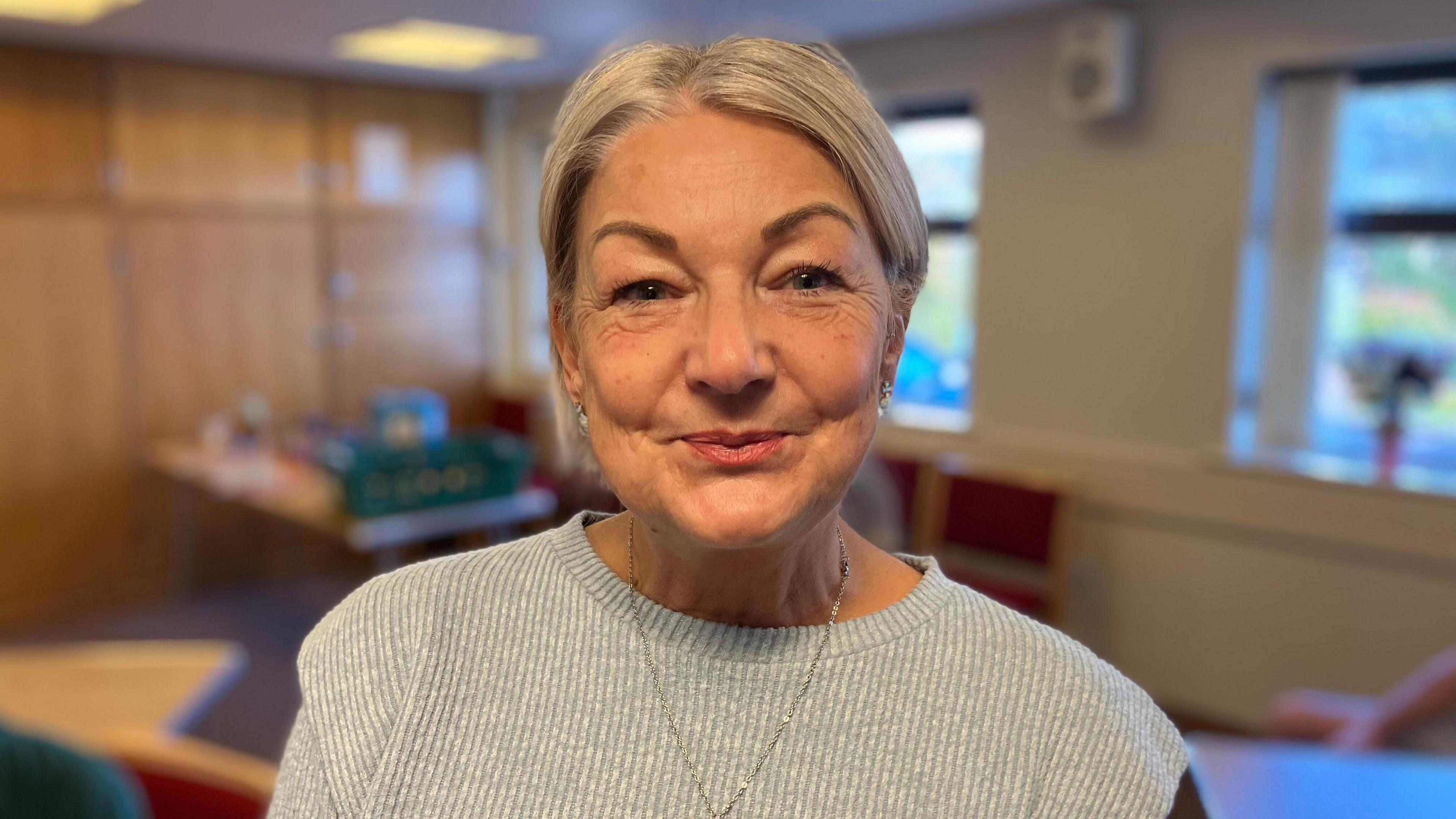 A women with blonde hair tied up, she is wearing a grey ribbed jumper whilst looking at the camera smiling.