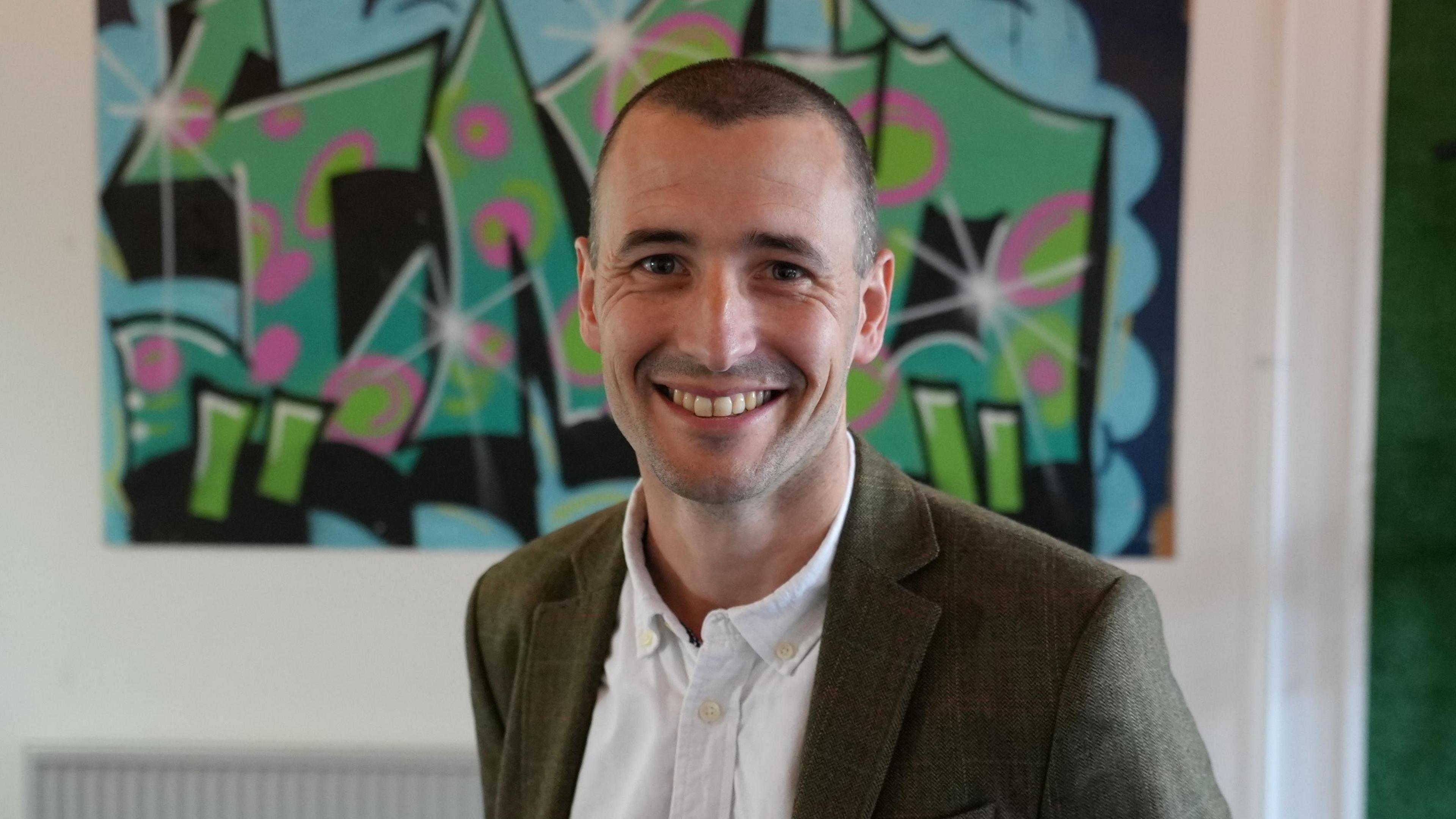 Ben Drabble has short hair and is wearing a green blazer over a white shirt. Behind him is a graffiti-style mural.