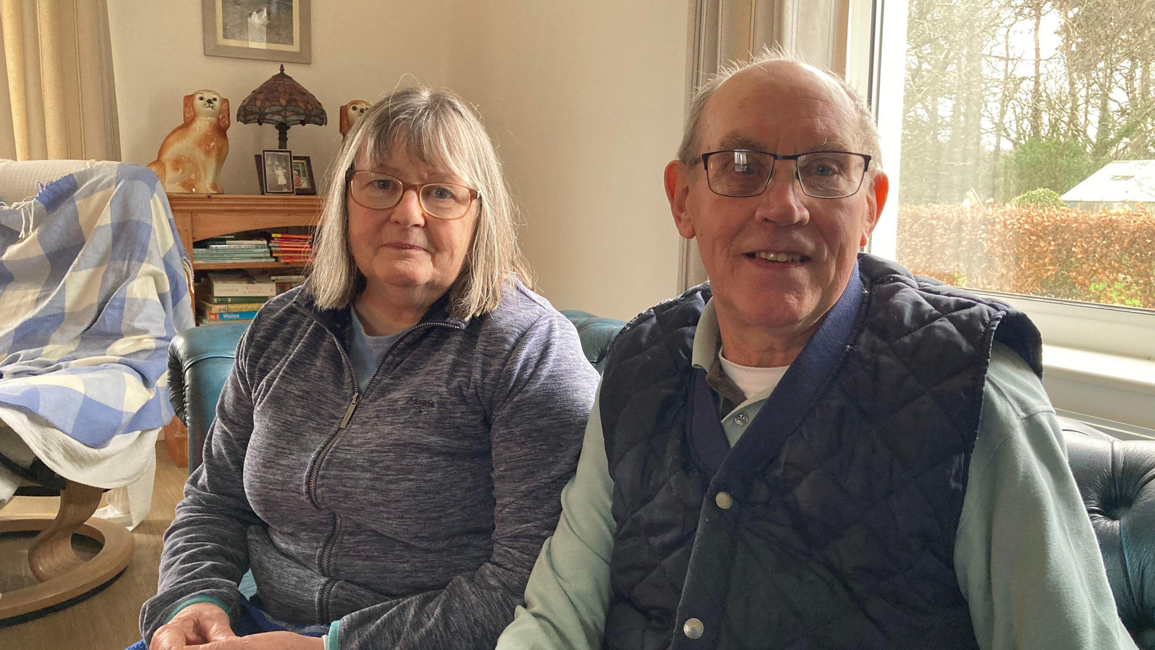 77-year-old Ronnie (right) and 71-year-old Barbara (left) sit in their home on the sofa. Barbara has a grey bob and full fringe and wears glasses and a zip up grey jacket, she looks blankly into the camera. Ronnie is bald and also has glasses on, he wears a navy quilted gilet and smiles into the camera. 