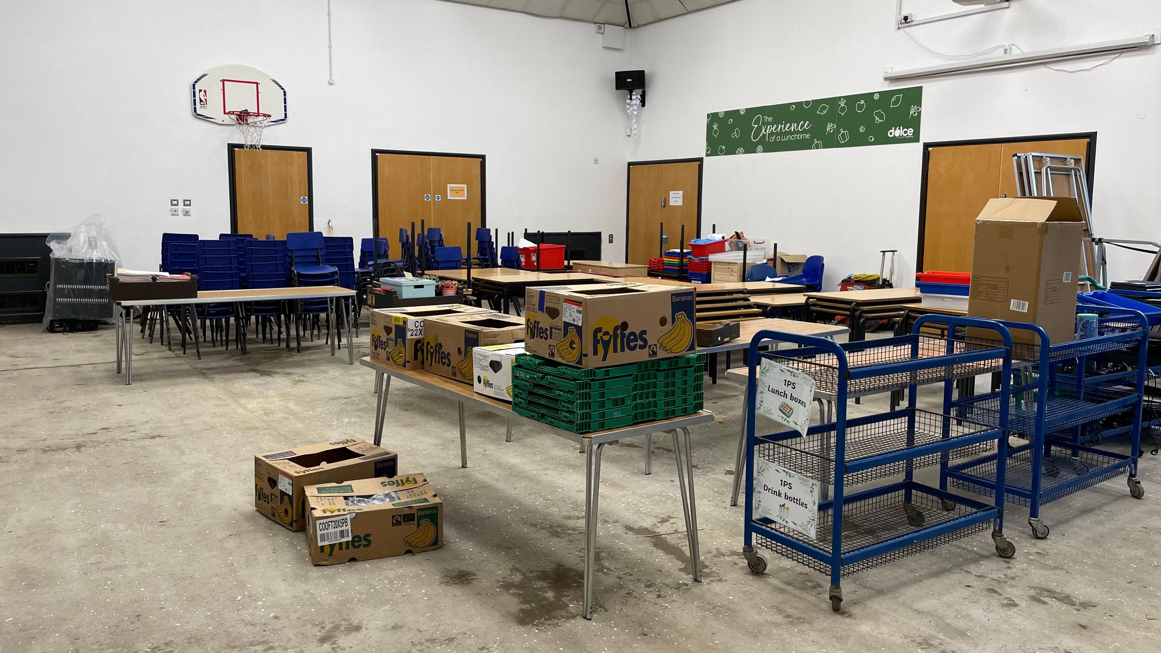 A school hall - a basketball net can be seen at one end, while they are tables and chairs stacked up on the floor