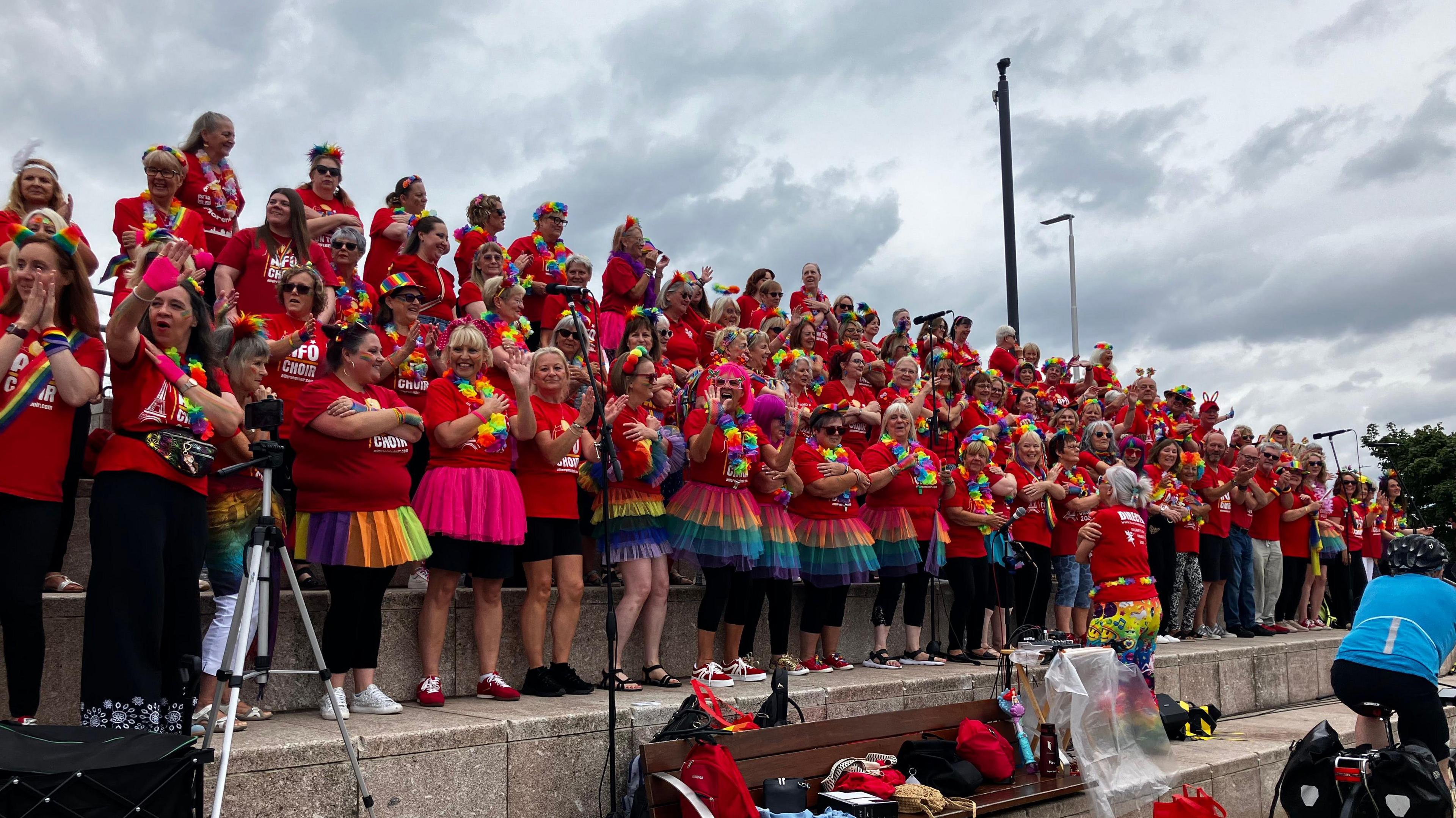 Dozens of singers in red T-shirts, standing in four tiered rows. Some of them are wearing rainbow tutu skirts. They are doing arm movements as they sing.