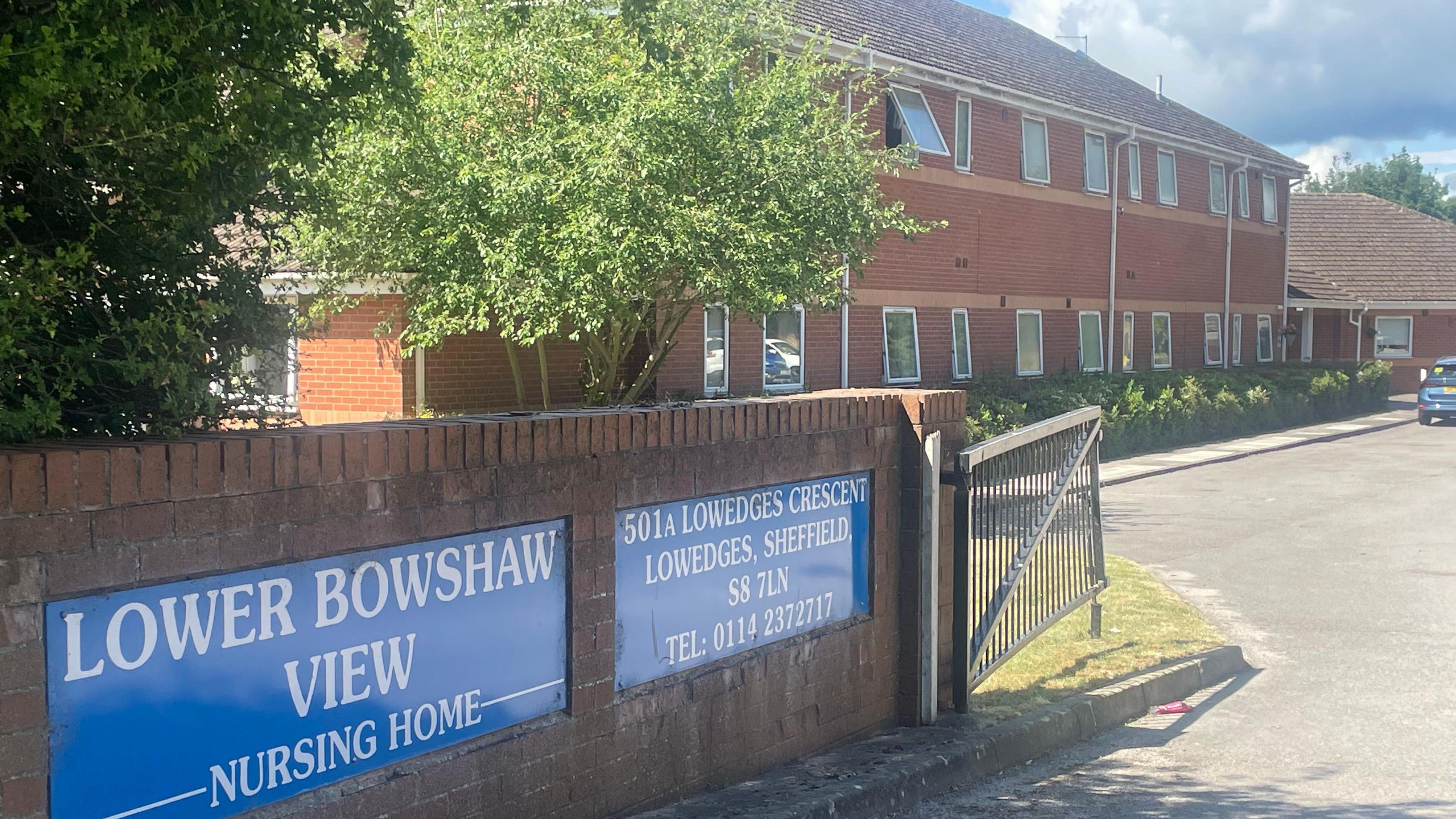 Lower Bowshaw View from its front gate. The main building is made from red brick, and the name of the home is printed on a blue sign next to the gate.
