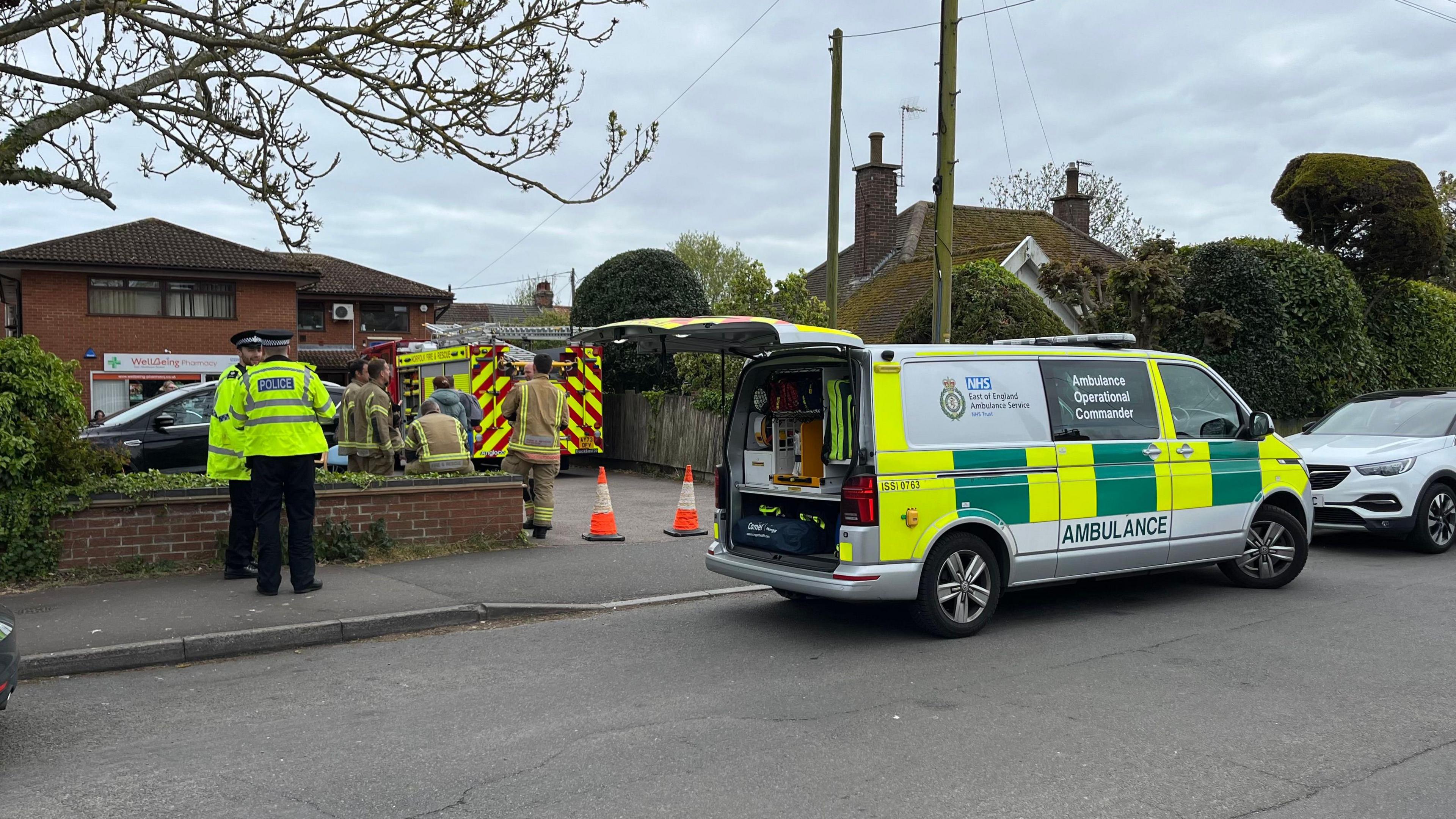 An Ambulance Operational Command vehicle is parked on the road outside the surgery with its boot open. Police officers stand nearby, there are orange cones blocking the entrance and a fire crew and engine in the car park.