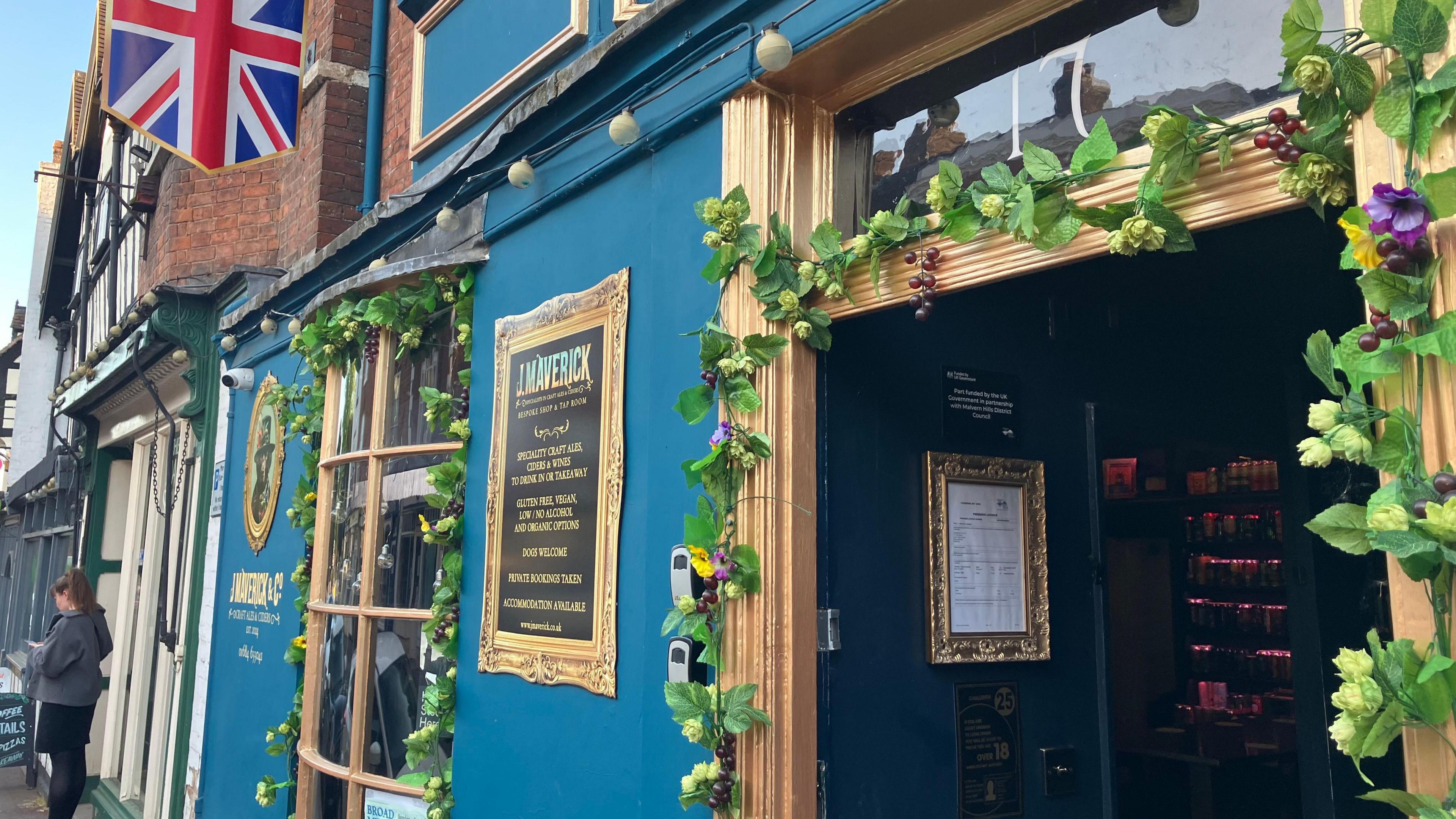 The outside of the pub which is painted blue and has a floral garland around the door. There are signs on the outside wall and inside the entrance. through the door, rows of drinks can be seen. The building has a bay window, which is also decorated, and there is a flag above the tap room. Further along the high street, a woman is standing underneath a black and white building.