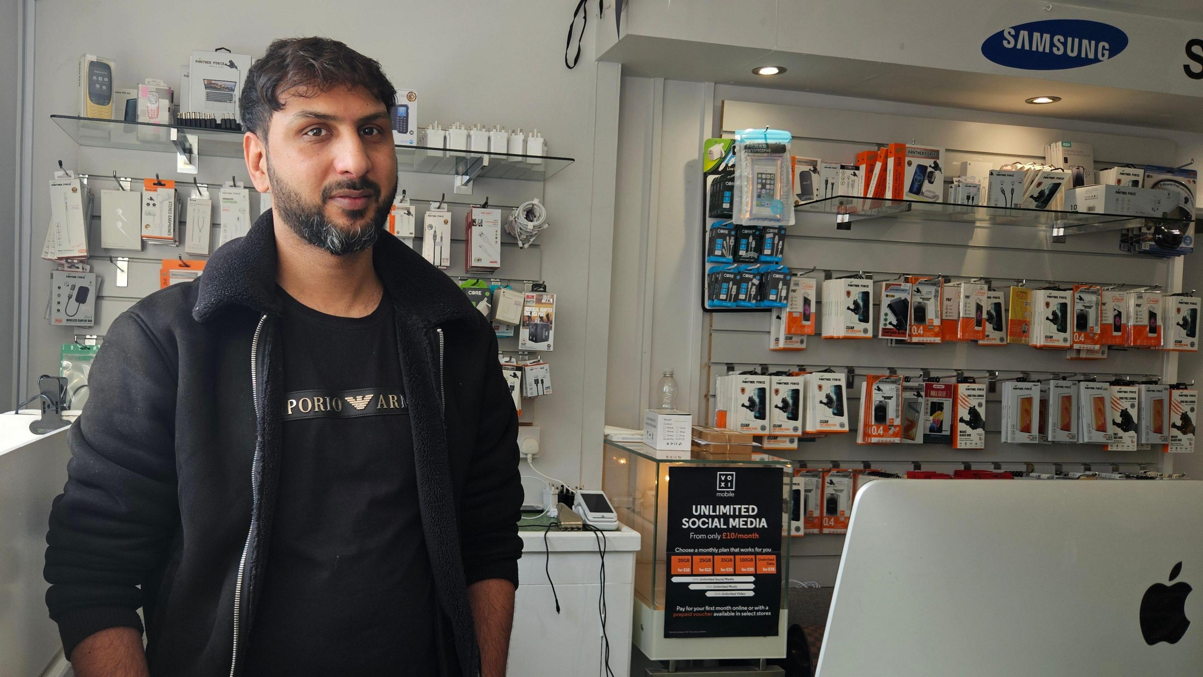 A young man of South Asian heritage stands behind a shop counter. On the wall behind him are many rows of items like mobile phone accessories and other electronics
