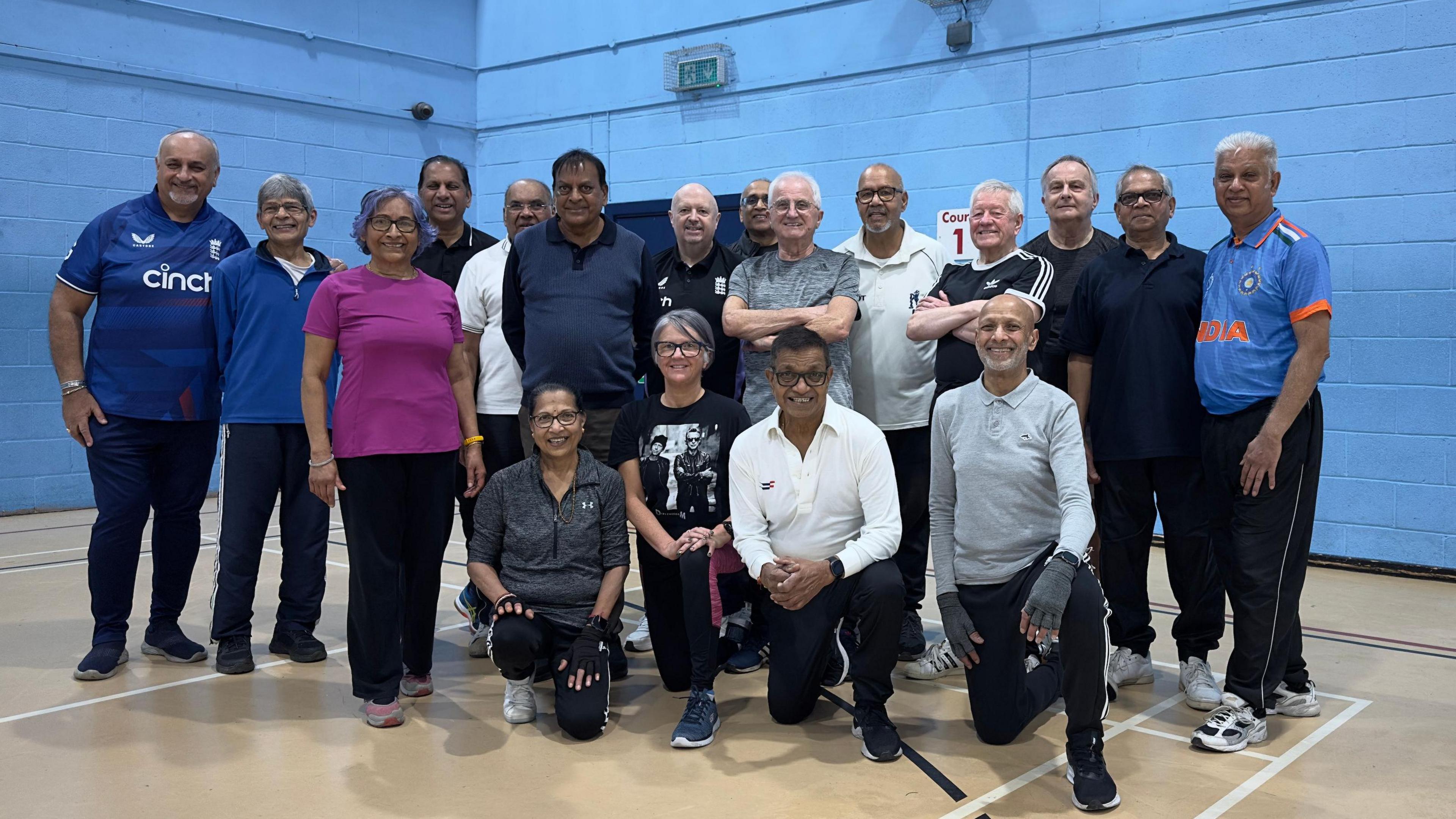 Members of the walking cricket club posing in sports wear in a sports hall standing against a blue-painted brick wall. 