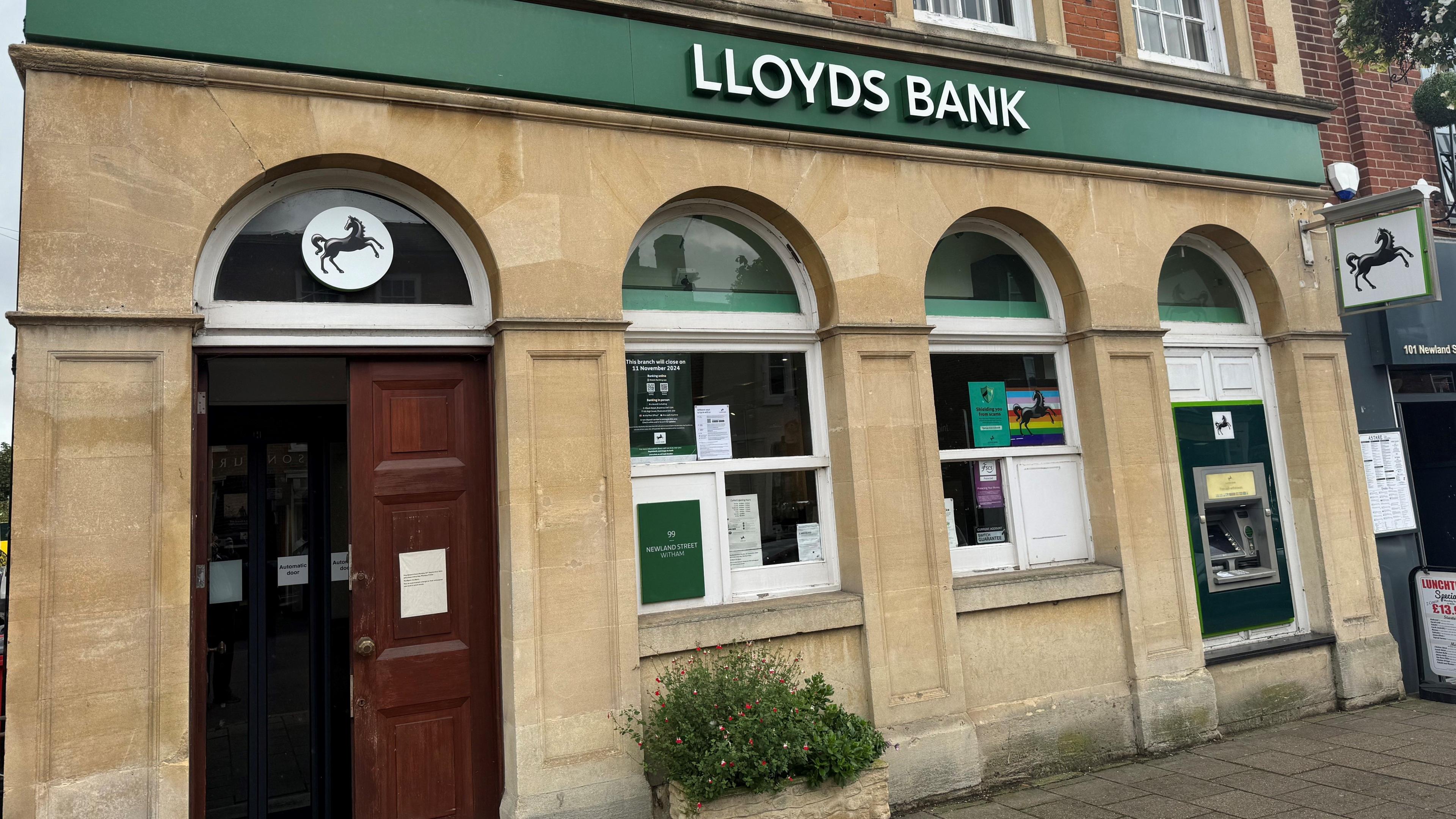 Outside a branch of Lloyds Bank. It is brick building with a brown wooden door, arched windows and a green banner above with the words "LLOYDS BANK".