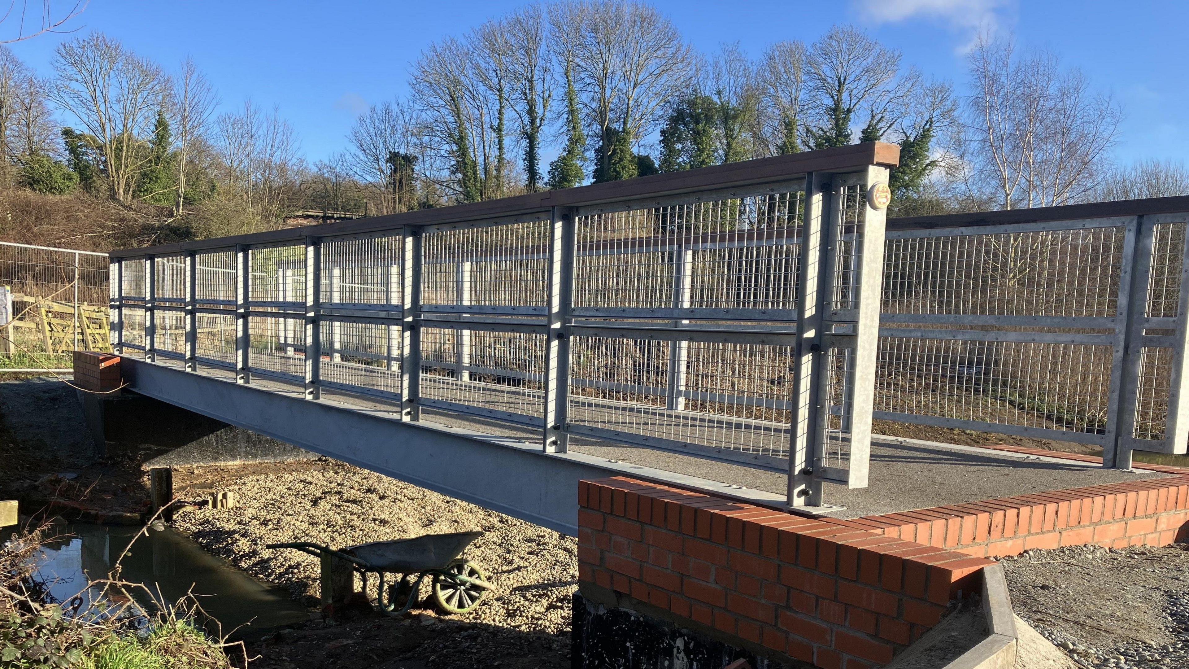 A new footbridge with grey metal railings and a red brick foundation spans a stream with a wheelbarrow underneath placed on shingle  