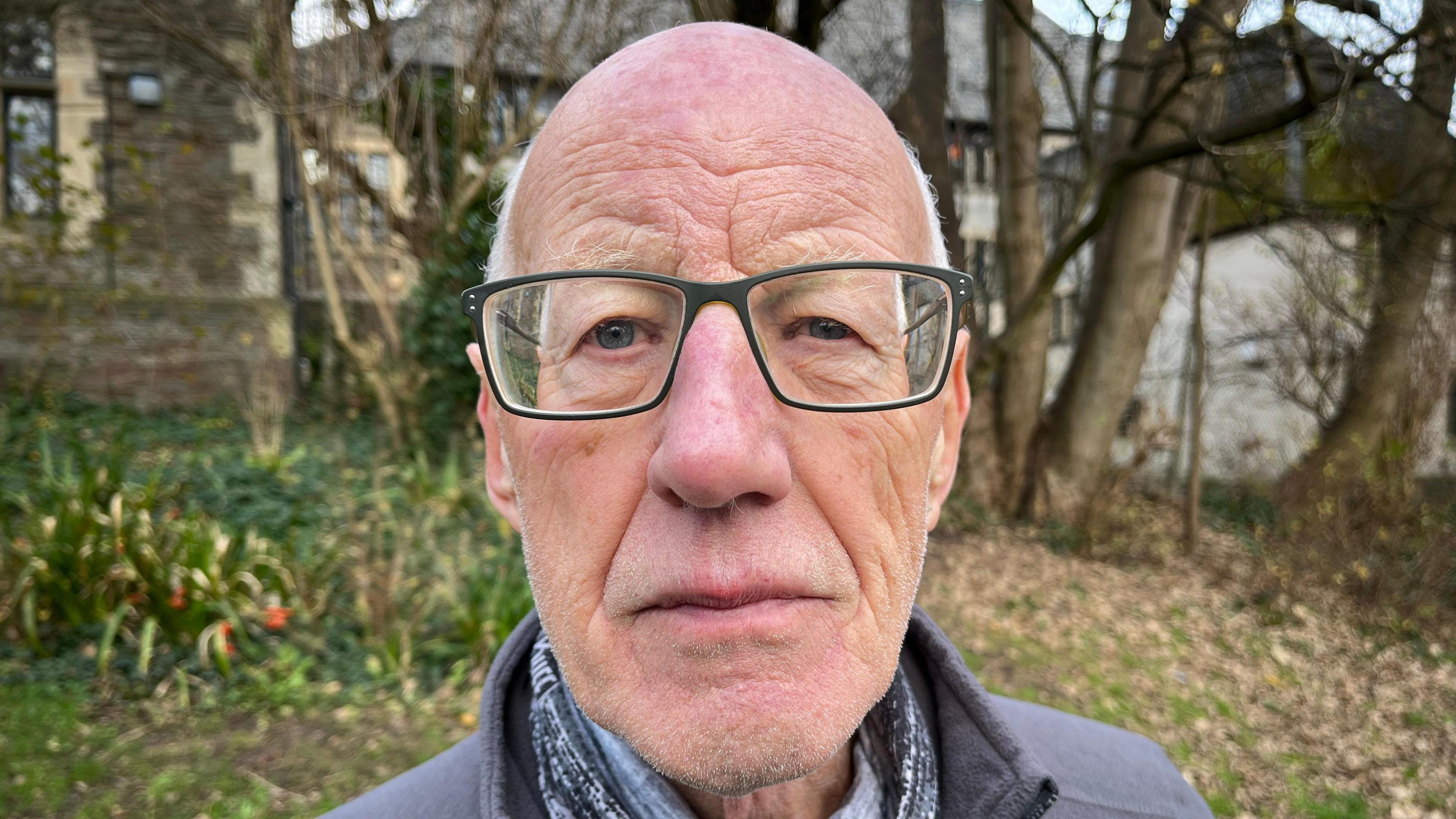 Mike Campbell looking directly into the camera. He is wearing glasses and a grey top. Behind him are some houses, and trees. 