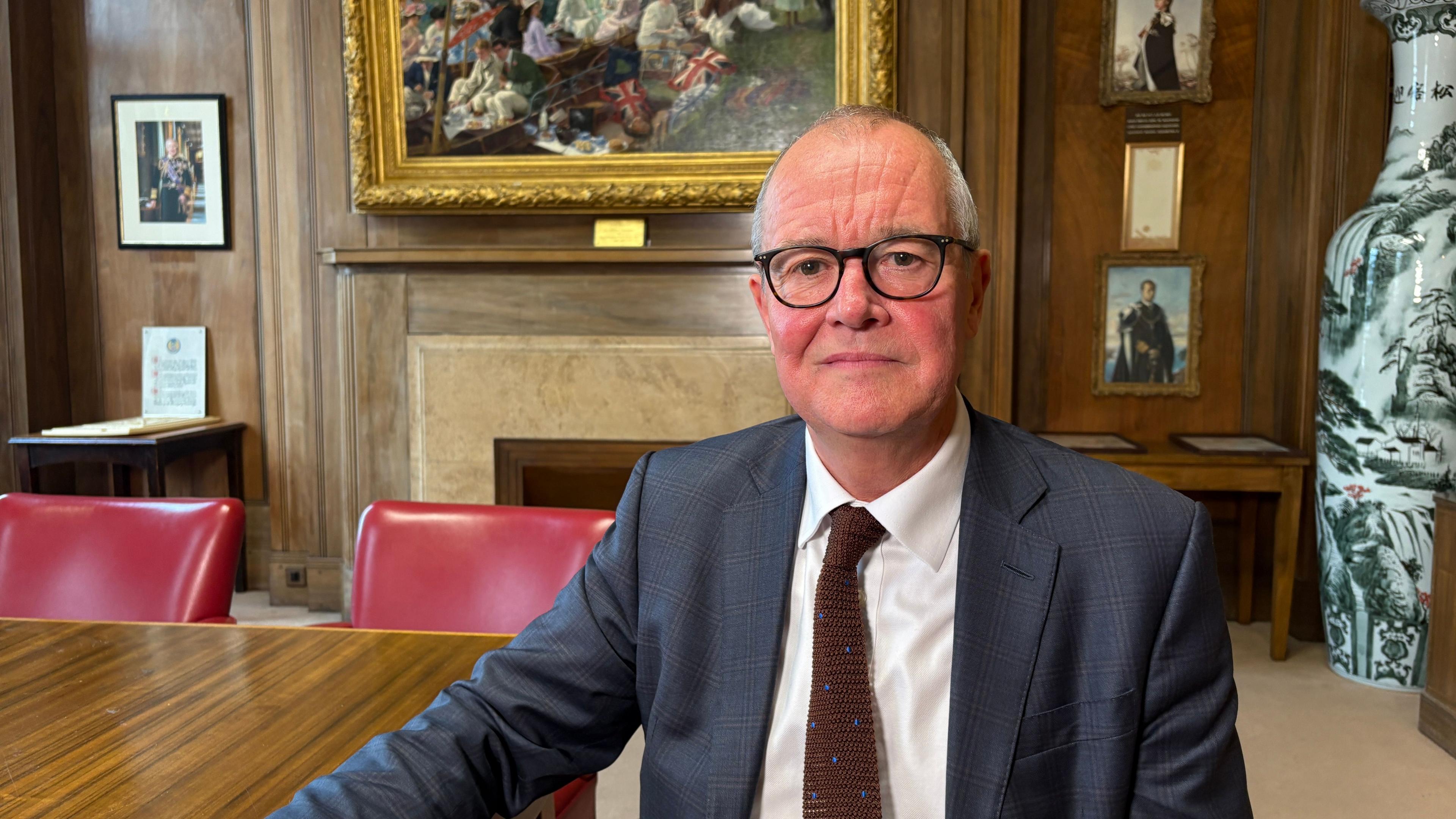 A formal room with wood-panelled walls and a polished wooden table surrounded by red leather chairs. Vallance is sitting at the table and looking at the camera. He is wearing a blue blazer, white shirt, red tie and black framed glasses. Behind him is a fireplace with a stone mantel, above which hangs a large framed painting of a historical scene.