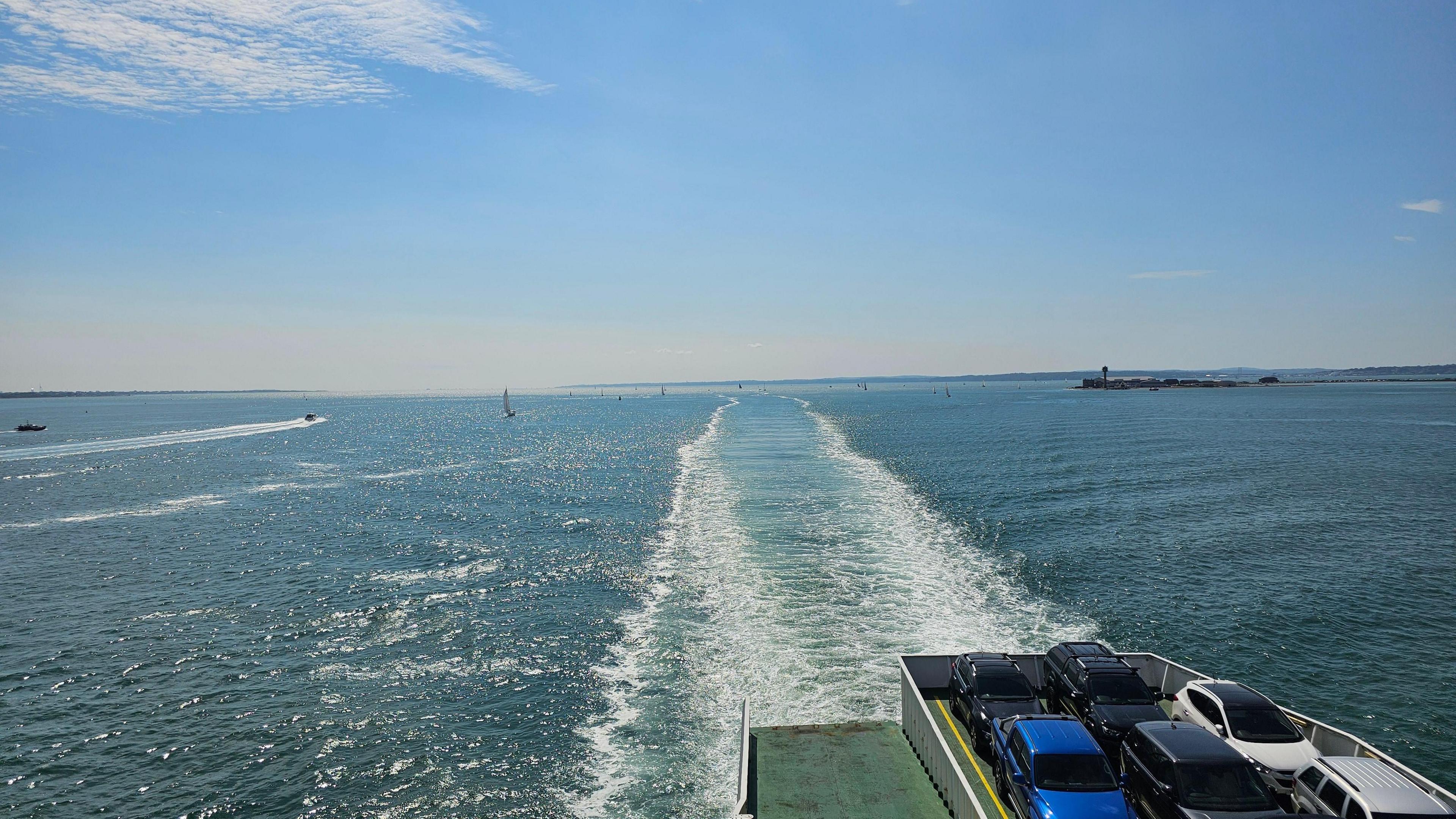 A small section of the rear of a car ferry can be seen in the foreground and the boat's wake can be seen behind it in the water. It is a sunny day with some small wispy clouds in the sky.