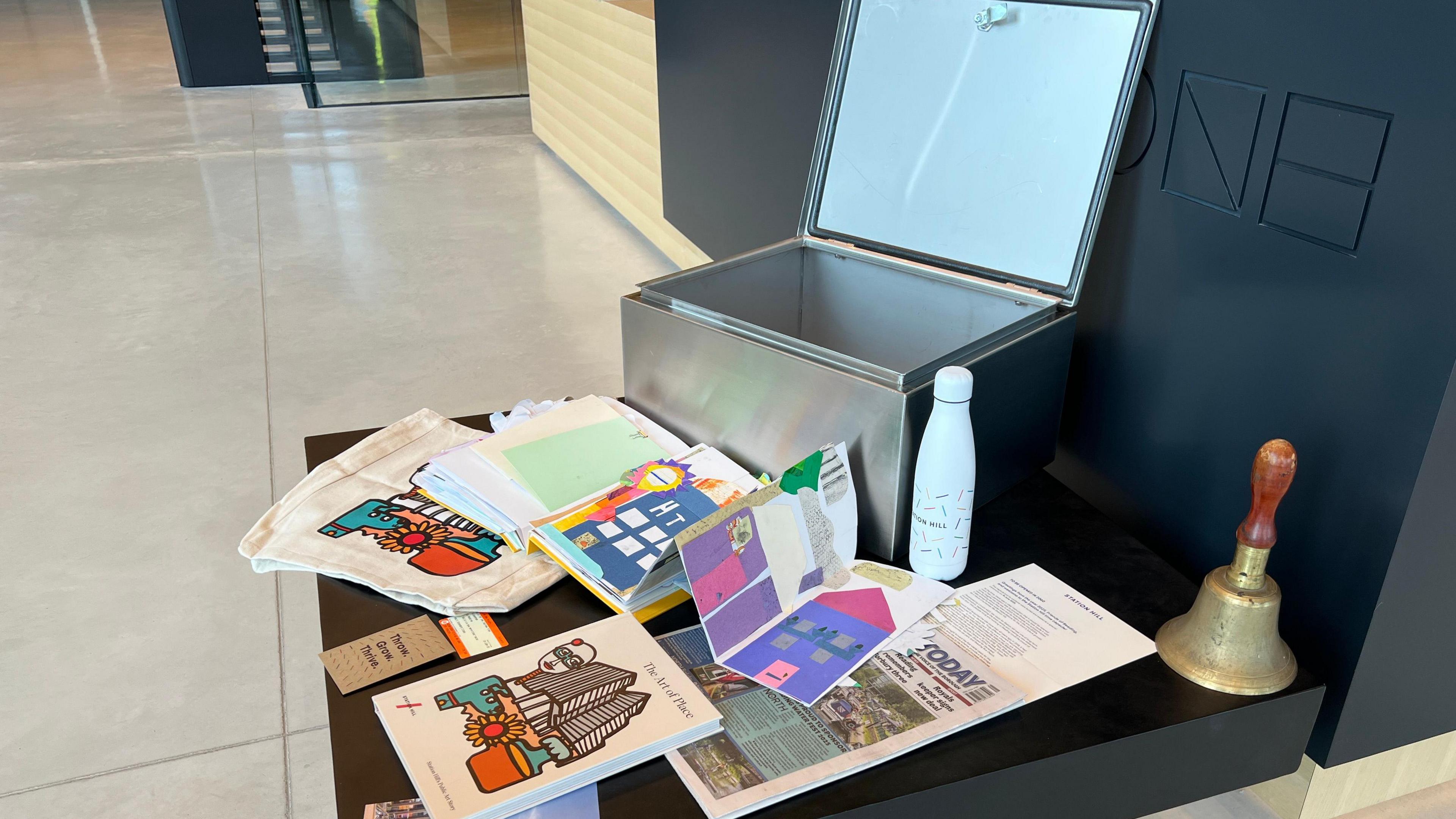The picture shows the capsule open and items around it which will be put inside to be buried. They include a copy of the local paper, a water bottle, pictures drawn by children, a book on art, a train ticket, and a bag.
