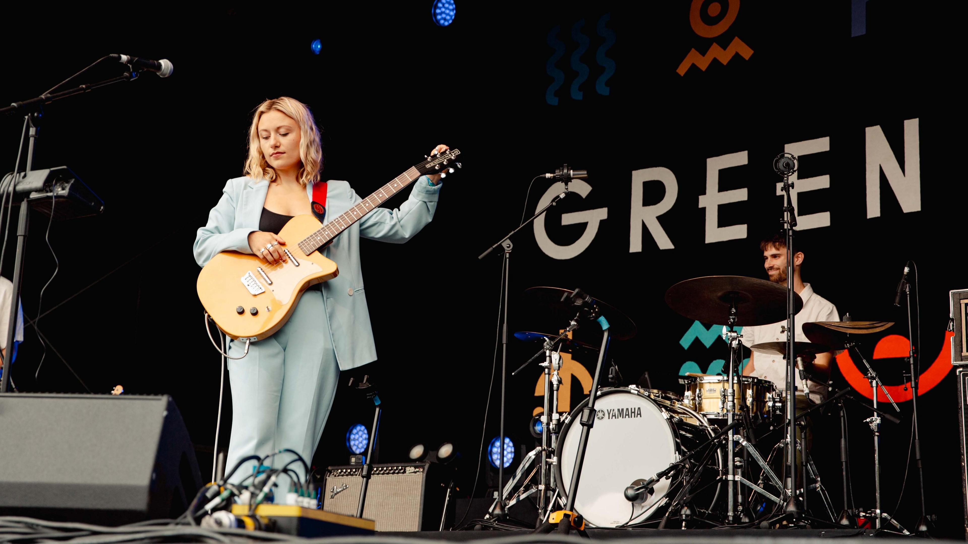 Gillie Rowland, holding a guitar and wearing a blue suit, performs at Green Man