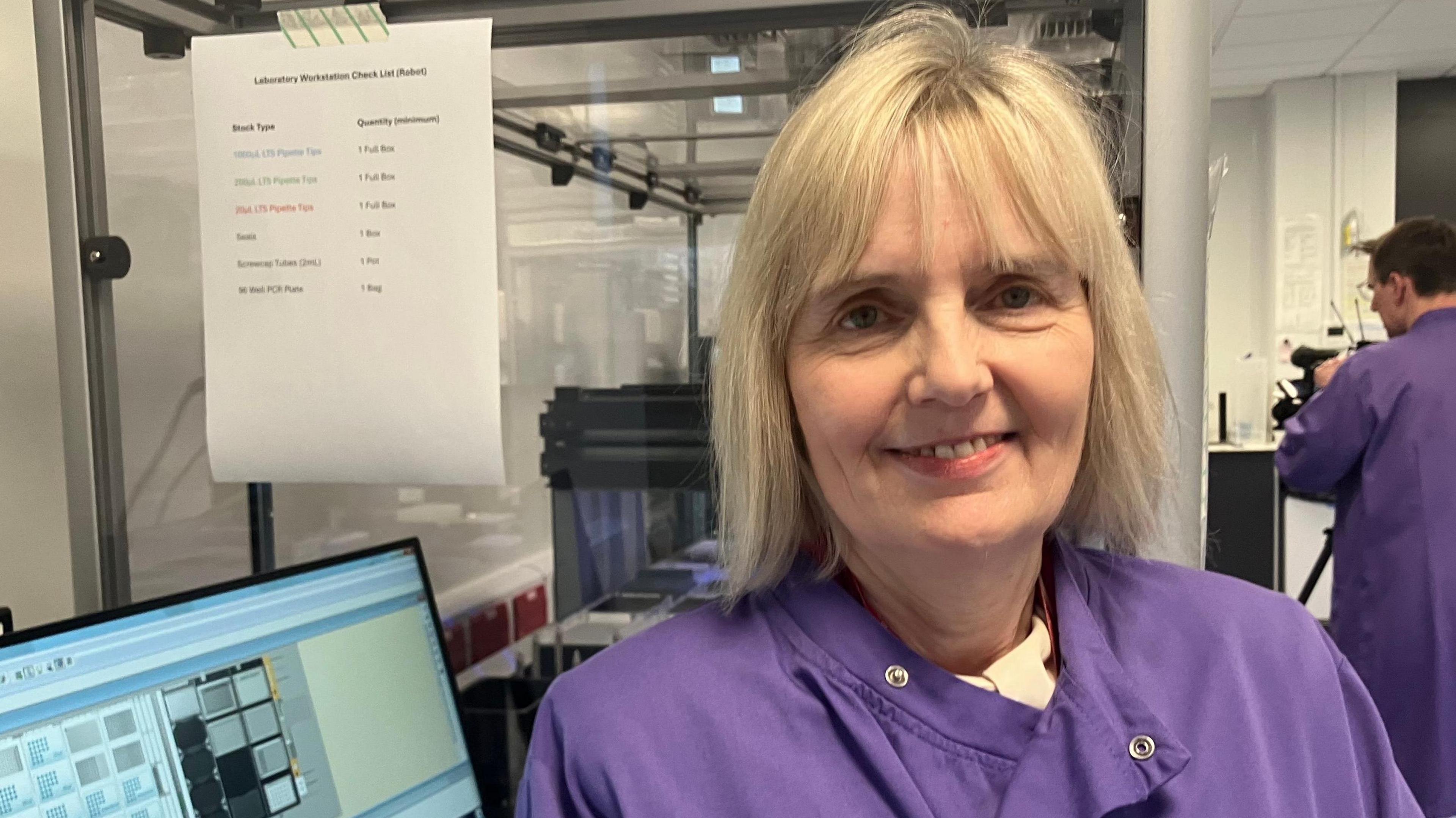 Sian yn gwisgo cot labordy piws. Mae offer technegol tu ol iddi. 