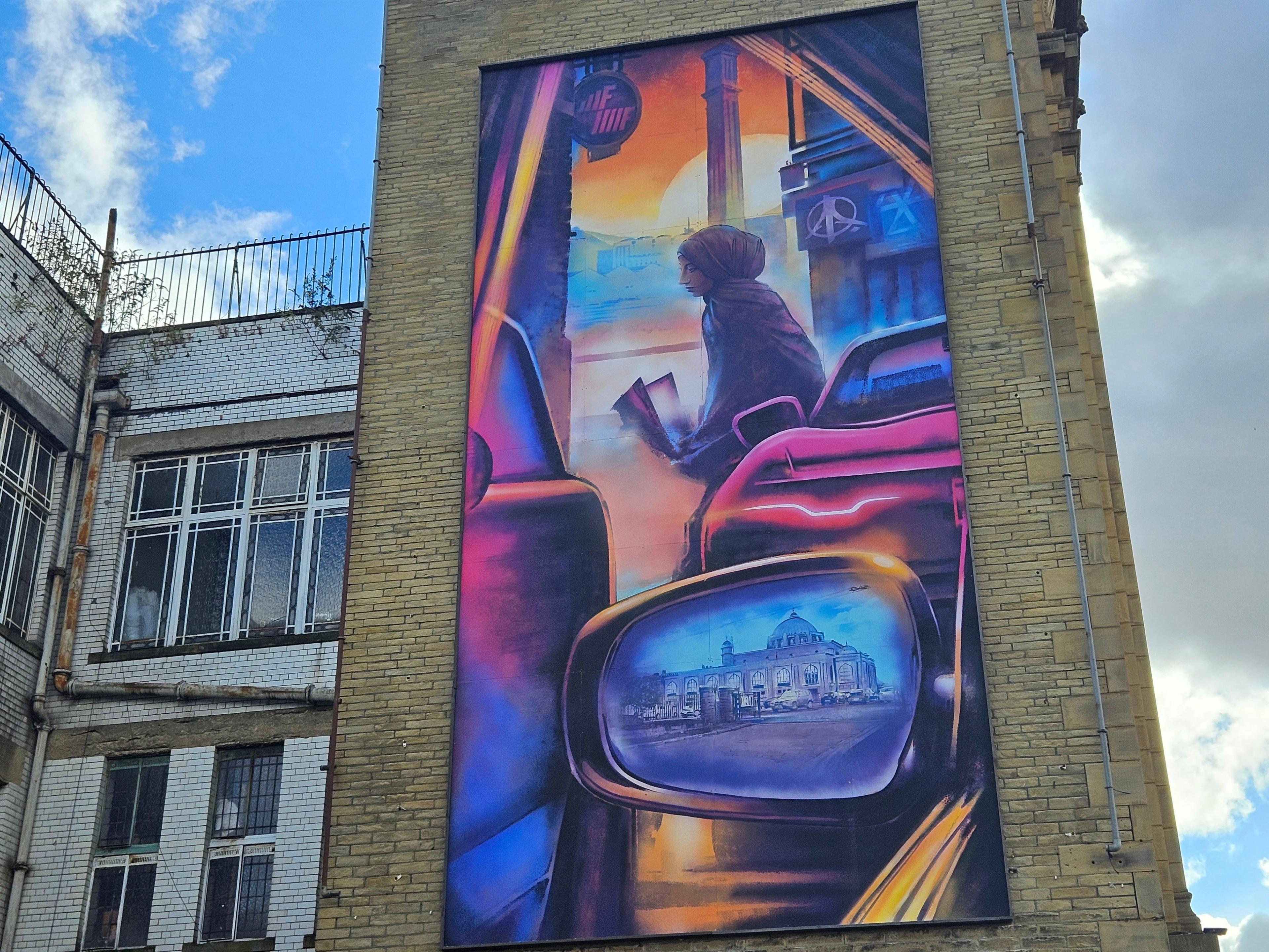 The mural depicts a young woman reading while she leans against a car with a cityscape reflected in her car’s wing mirror.