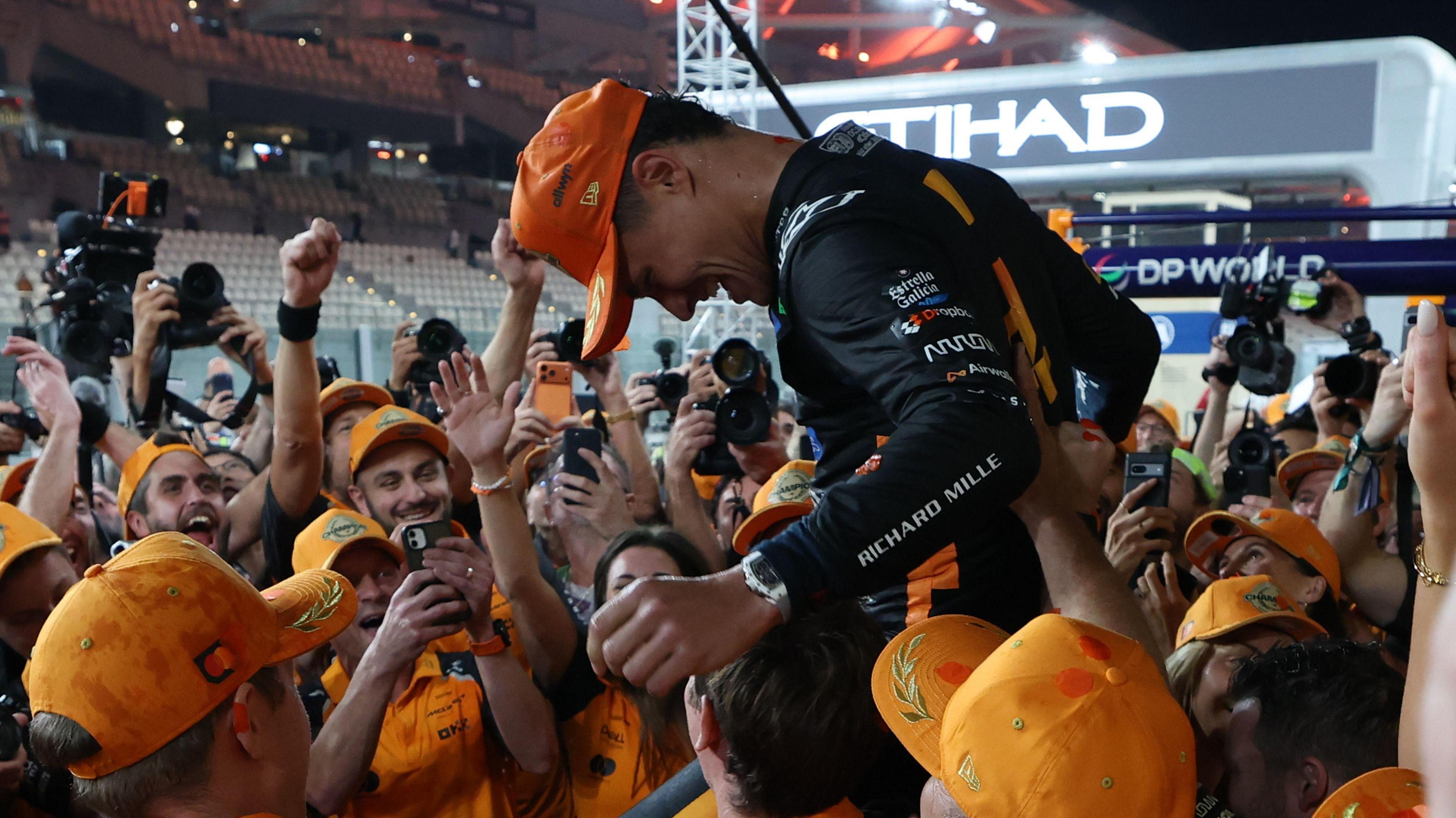 F1 driver Lando Norris celebrates his win by being held up by his team members and smiling